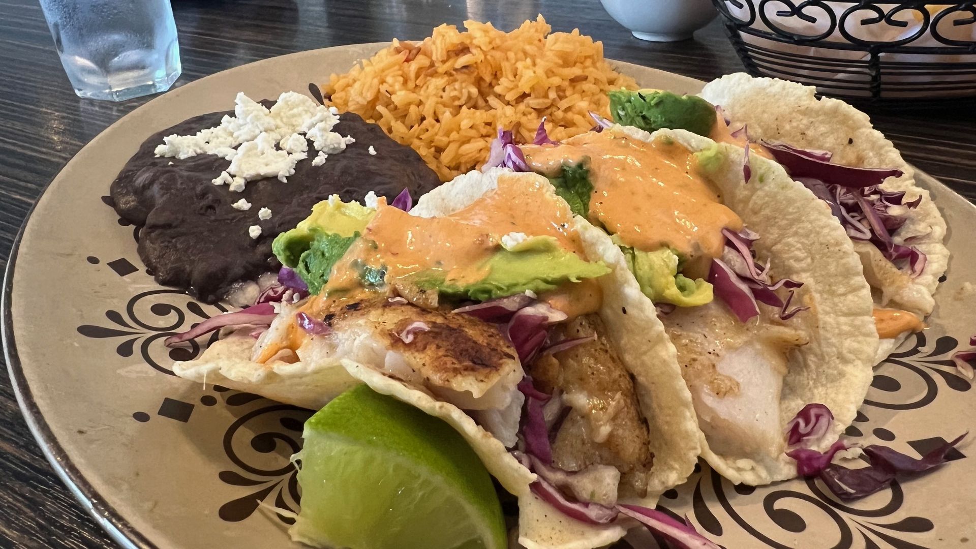 Plate with three fish tacos topped with avocado and orange sauce, served with black beans topped with cheese, Spanish rice, and a lime wedge on the side.