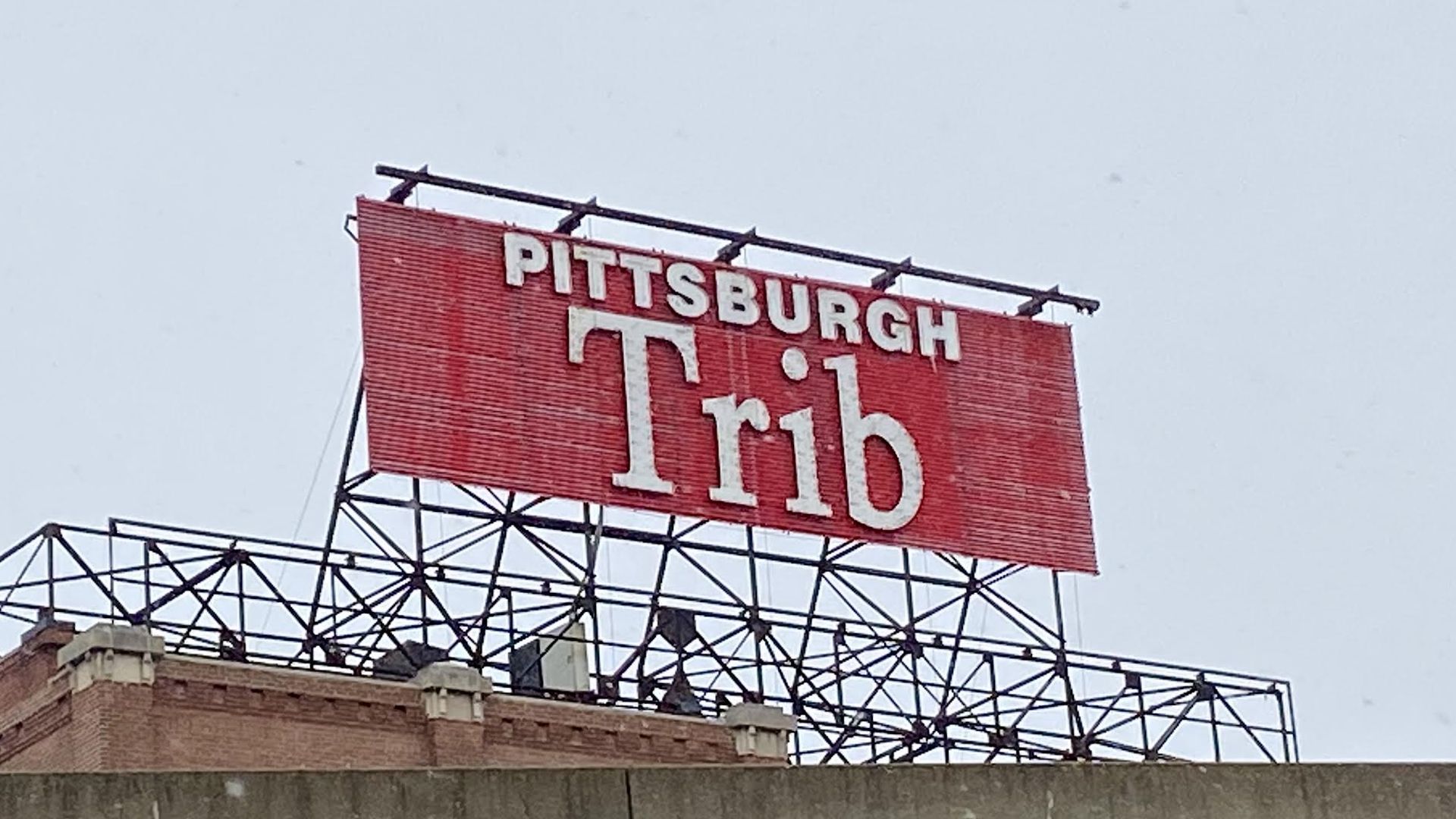 Large red rooftop sign reading "PITTSBURGH Trib" in white letters atop a brick building with metal framework against a gray sky.