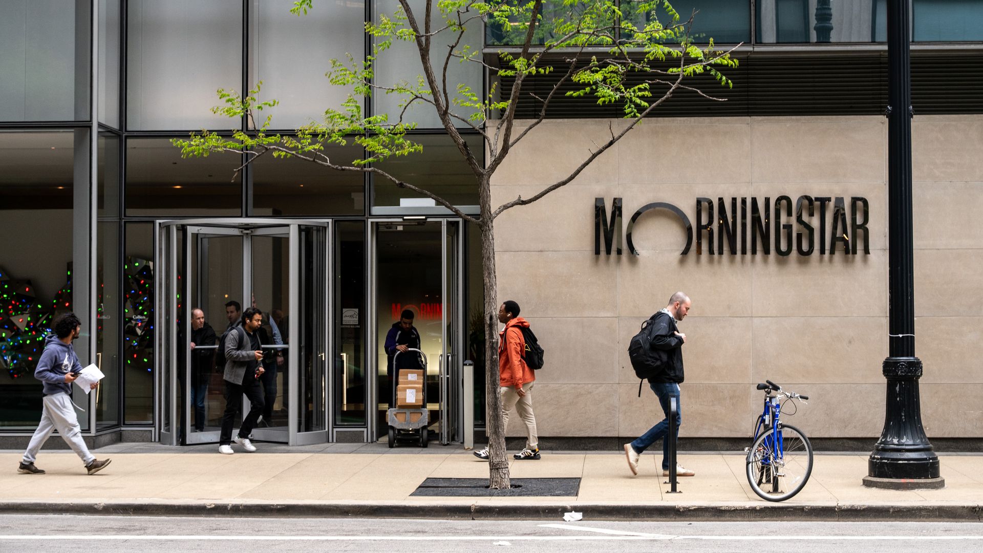 Urban storefront with a leafy tree in front, glass panels above, and a beige wall reading "MORNINGSTAR"; pedestrians walk by as a blue bicycle leans by a lamppost.
