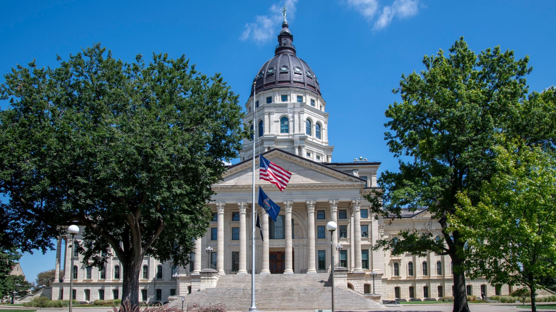 Kansas' state capitol building in Topeka with a dome and white columns, and in the foreground, green trees and a flag pole supporting the American flag and, below it, the Kansas state flag.