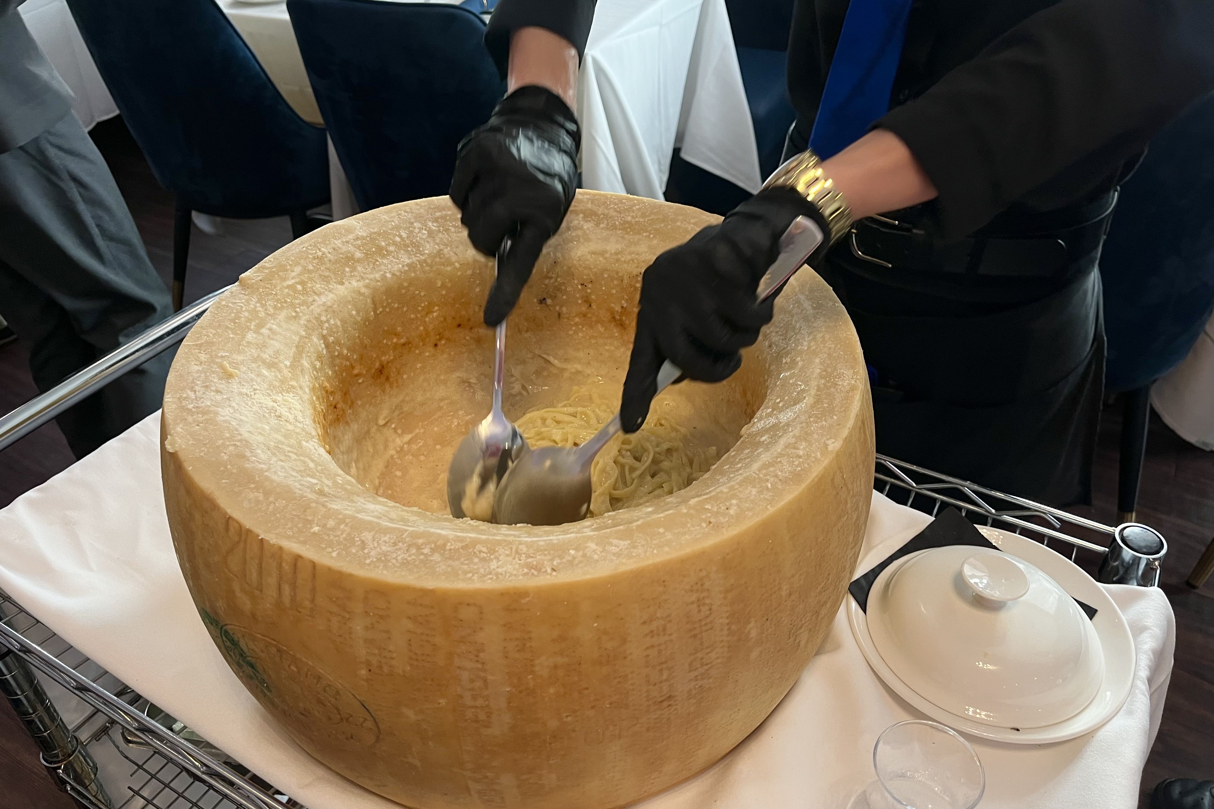 A person wearing black gloves is preparing pasta inside a hollowed-out wheel of Parmesan cheese. The large cheese wheel is placed on a cart with a white tablecloth. Nearby, there is a covered dish and a small glass.