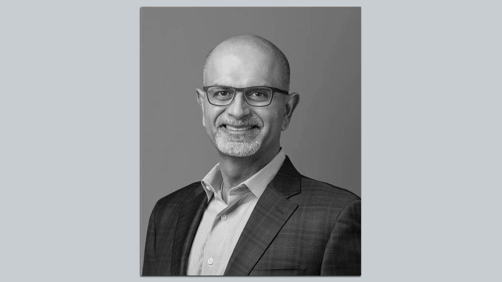 Black-and-white portrait of a smiling, balding man with glasses and a gray goatee, wearing a checkered suit jacket and light shirt, posed against a plain gray background.