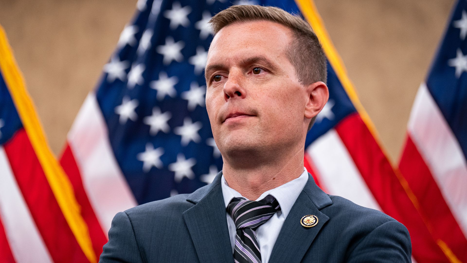Rep. Jared Golden (D-Maine), wearing a navy jacket, black and purple striped tie and a pale blue shirt, stares ahead in front of 3 US flags.