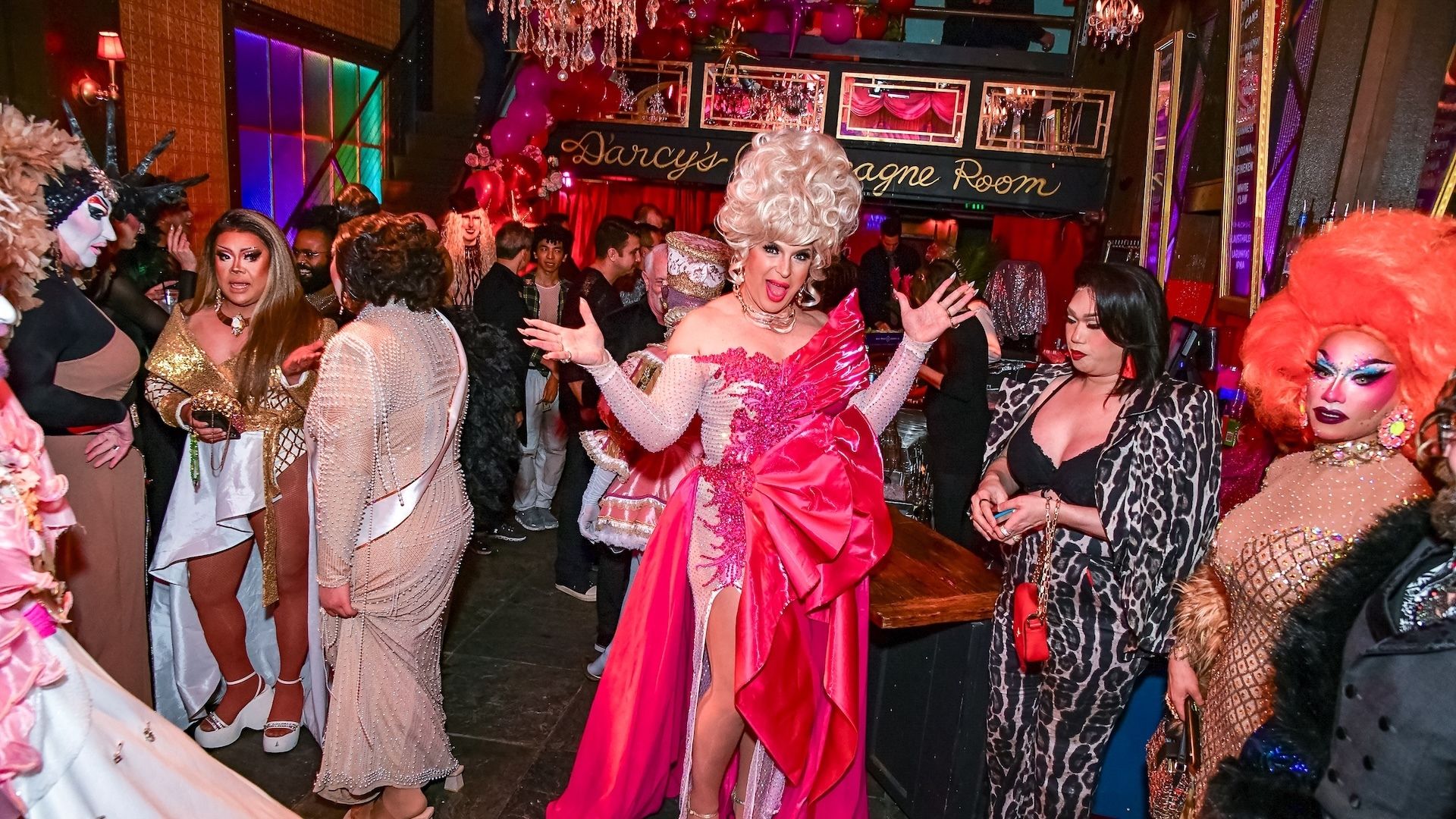 Lively drag show crowd in colorful, glamorous outfits and makeup, featuring a central figure in a vibrant pink dress and large blonde wig.