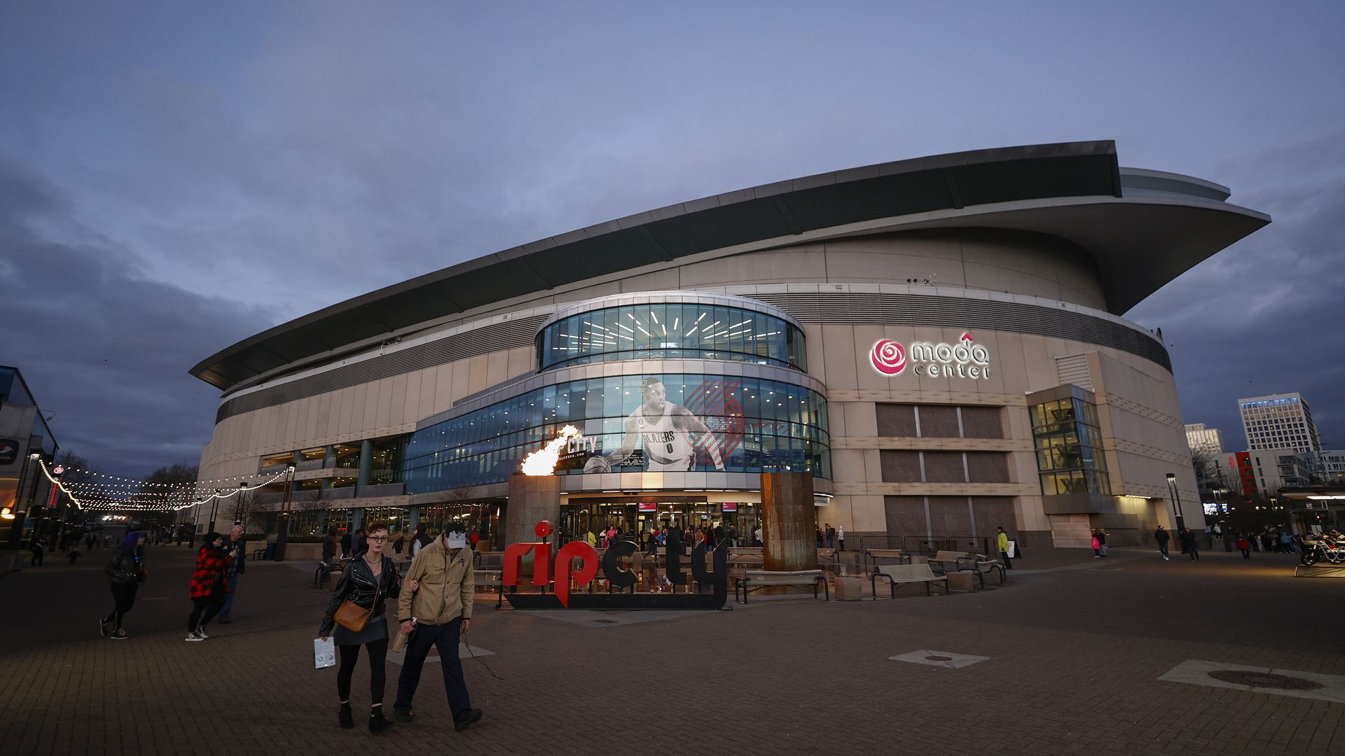 The exterior of Portland's Moda Center, a low, rounded building, with a video display of basketball players and a sign that says Rip City in front. 
