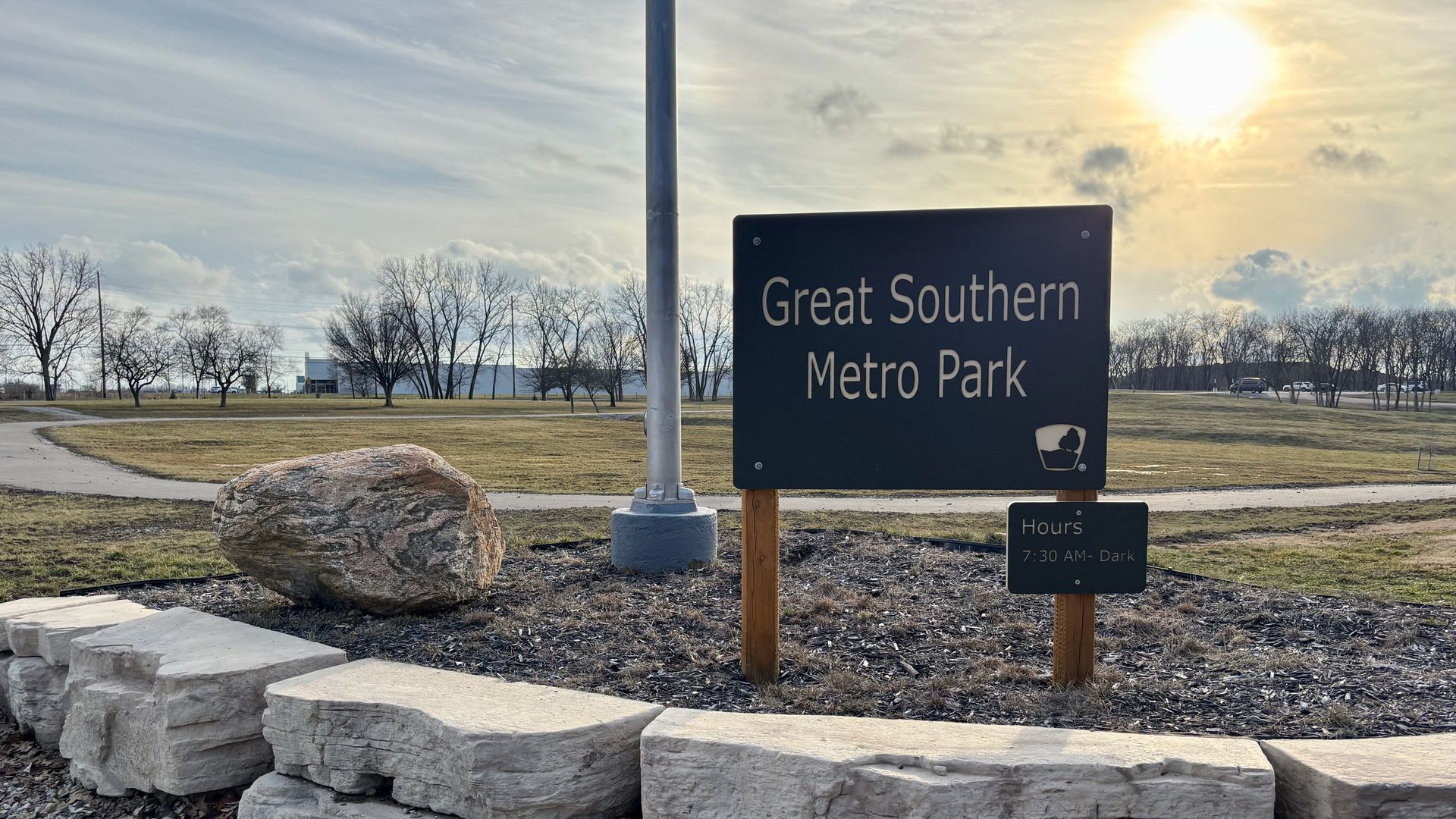 Sign for Great Southern Metro Park with park hours 7:30 AM to dark, surrounded by large light-colored stones, a boulder, bare trees, grassy field, and a partly cloudy sky with the sun low.