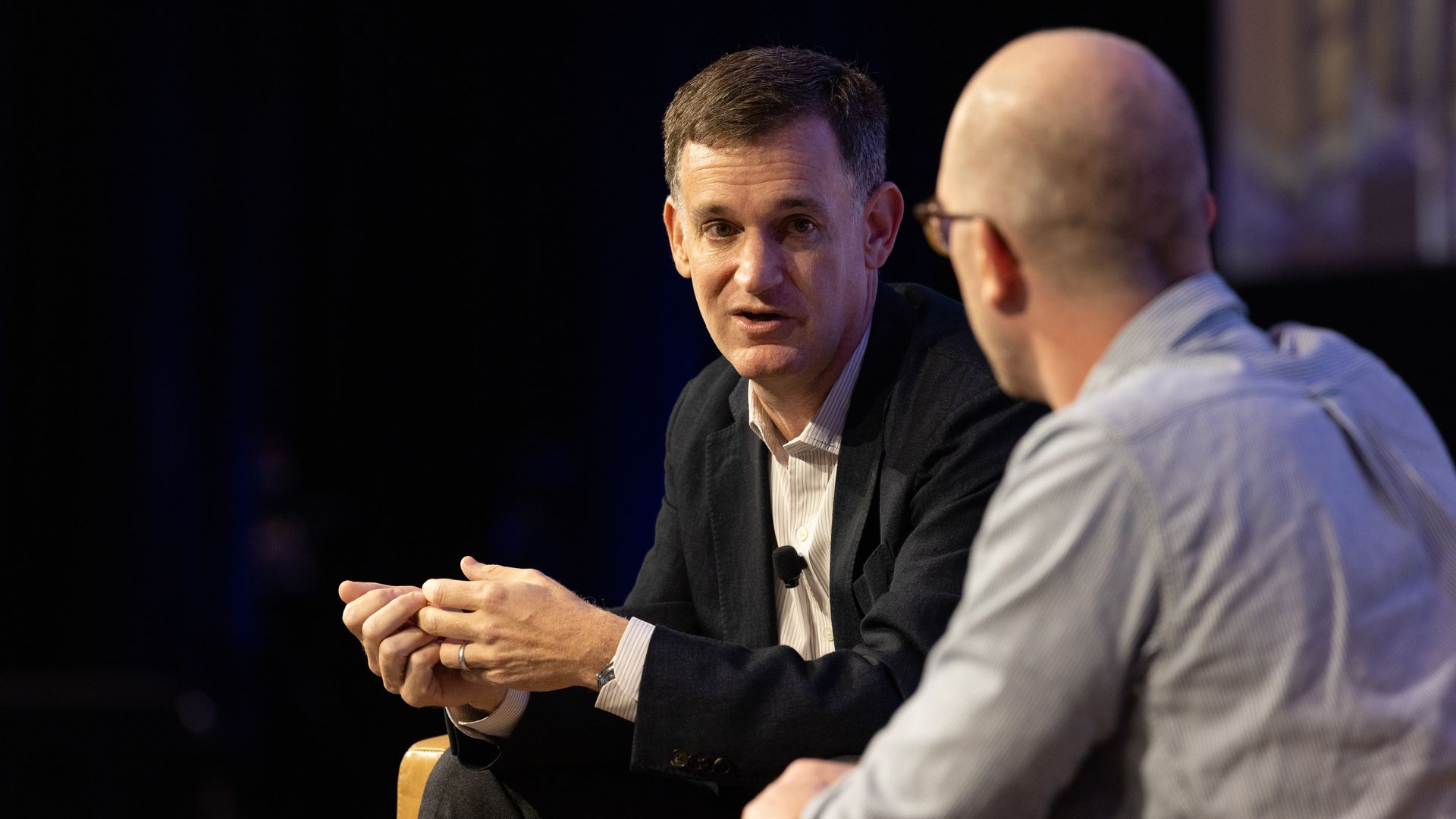 Arnold Ventures chair John Arnold, sitting at left in a dark sport coat, speaks on stage in a darkened ballroom, to a reporter sitting at right, facing away from the camera, in a blue striped shirt.