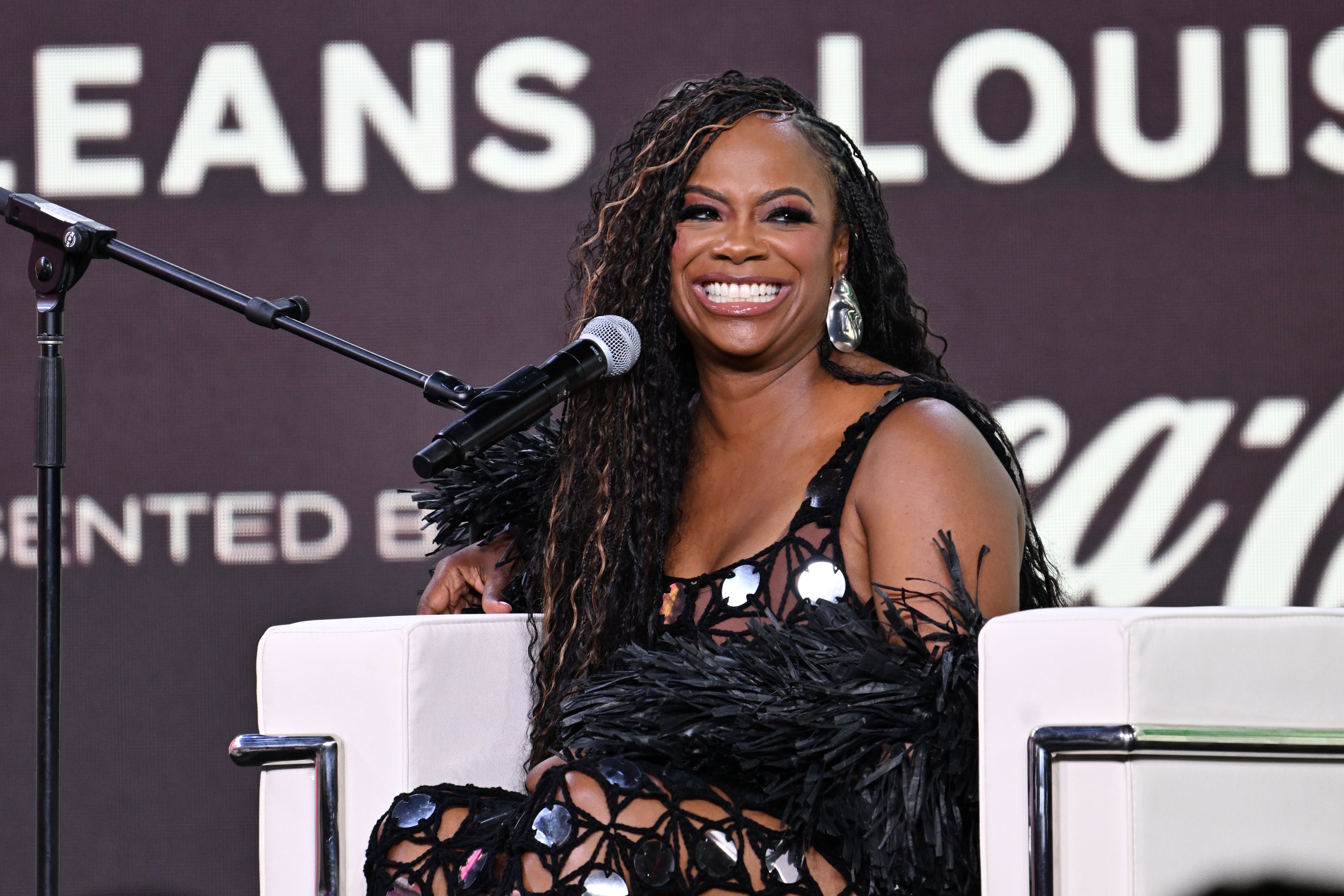 Smiling woman with long braided hair wearing a black dress with feathers and large silver earrings, sitting on a white chair in front of a microphone and dark background with white text.