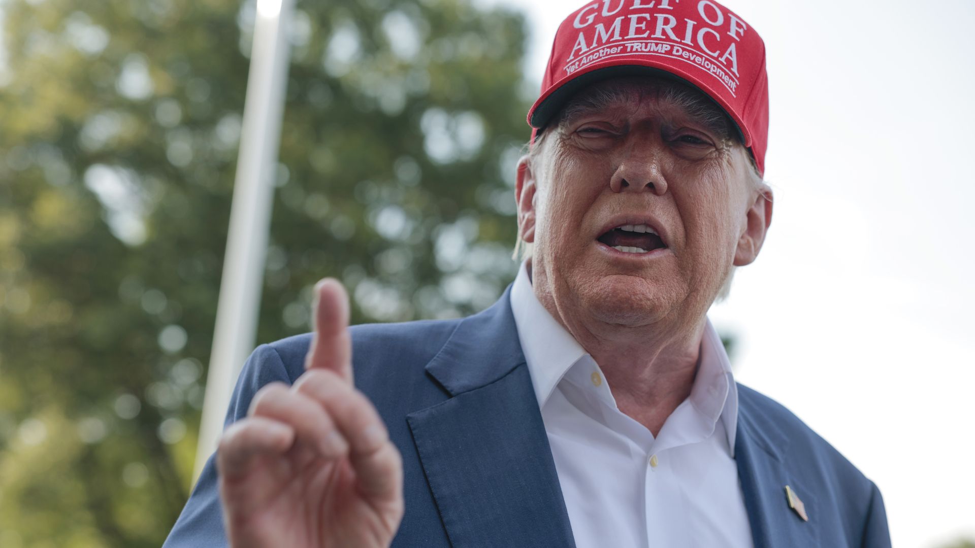 Donald Trump wearing a tieless blue suit and a red "GULF OF AMERICA" hat, raising a finger while standing in front of a tree and a flagpole.