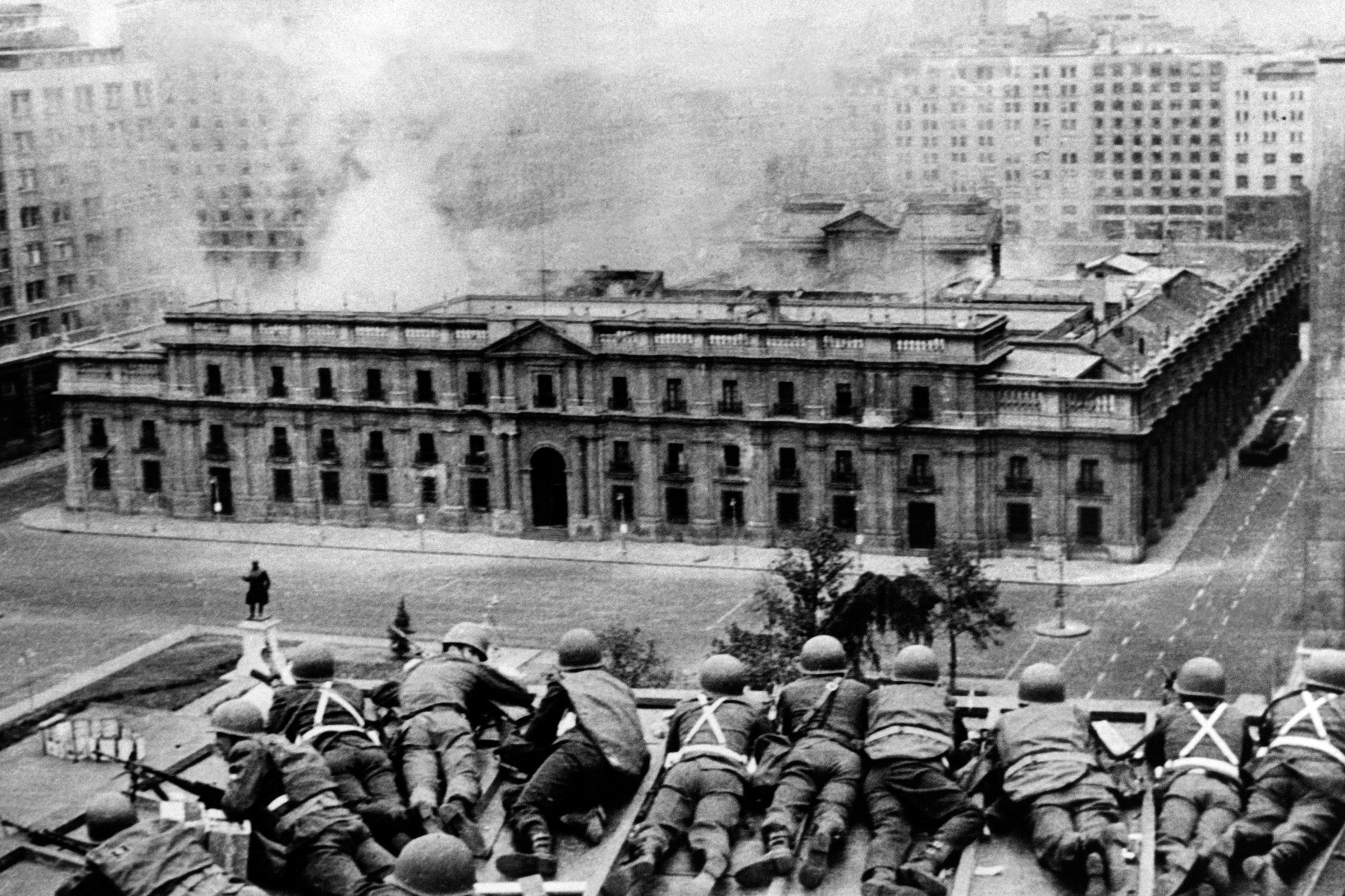 A black and white photo shows soldiers lying on their stomaches while aiming weapons at Chile's  Moneda palace, which has smoke coming out, after a coup on Sept. 11, 1973