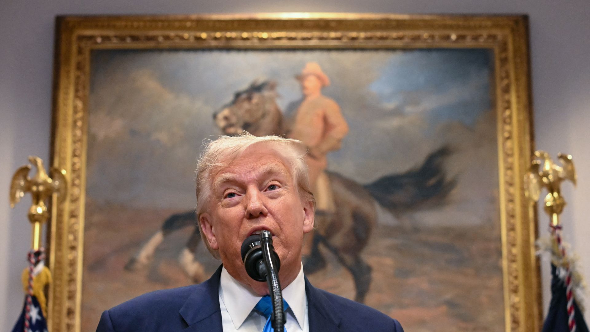 President Trump, in a blue suit and blue tie, speaks into a microphone in front of a framed painting of a person on a horse, with two gold eagle decorations and American flags on each side.