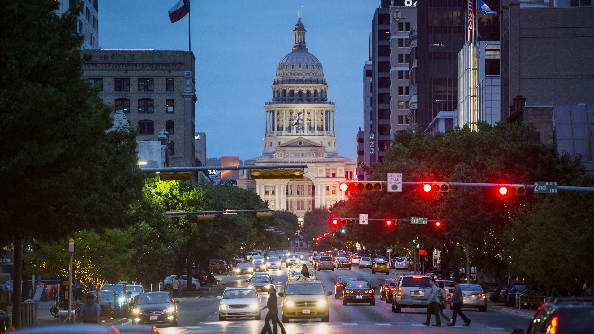 The Texas State Capitol building stands past cars and pedestrians crossing the street at dusk in Austin