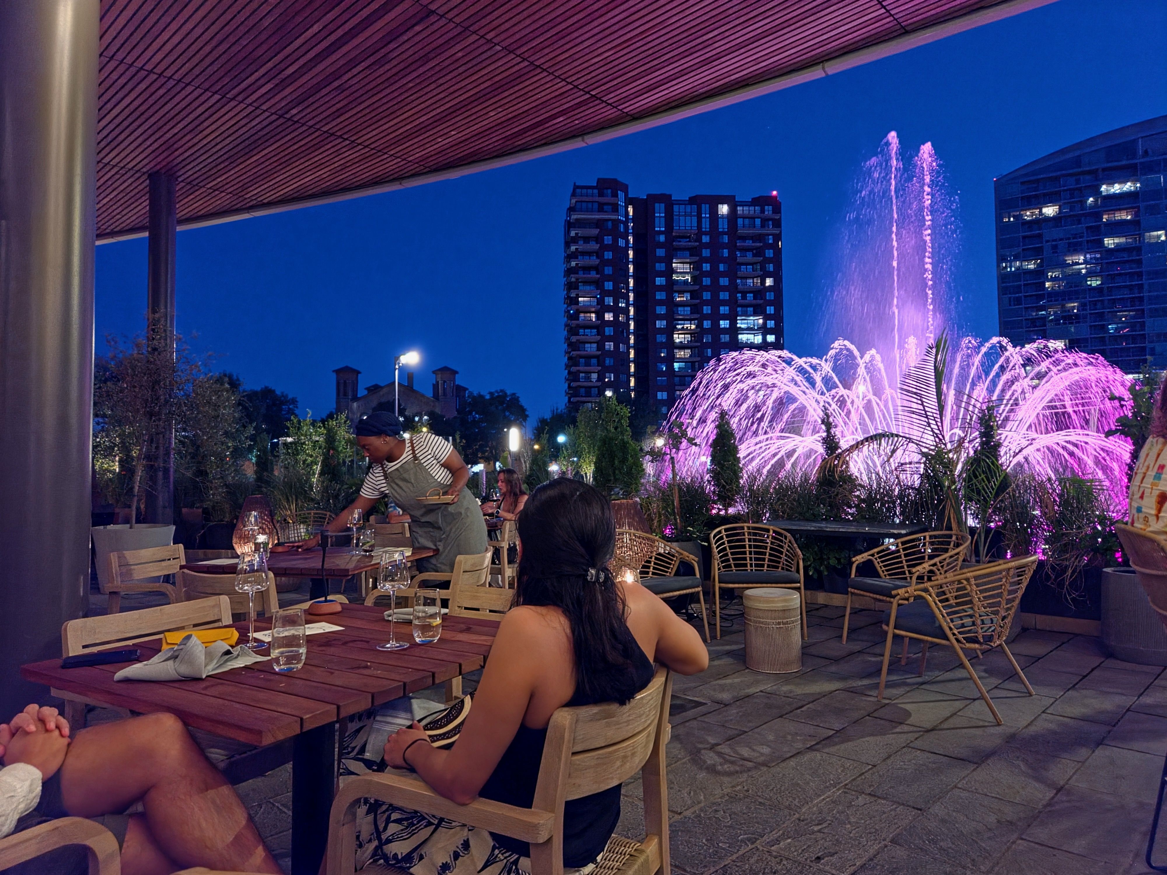 Nighttime outdoor restaurant scene with a purple-lit fountain and modern buildings in the background. Diners sit at wooden tables; a waiter in an apron serves guests under a covered patio.