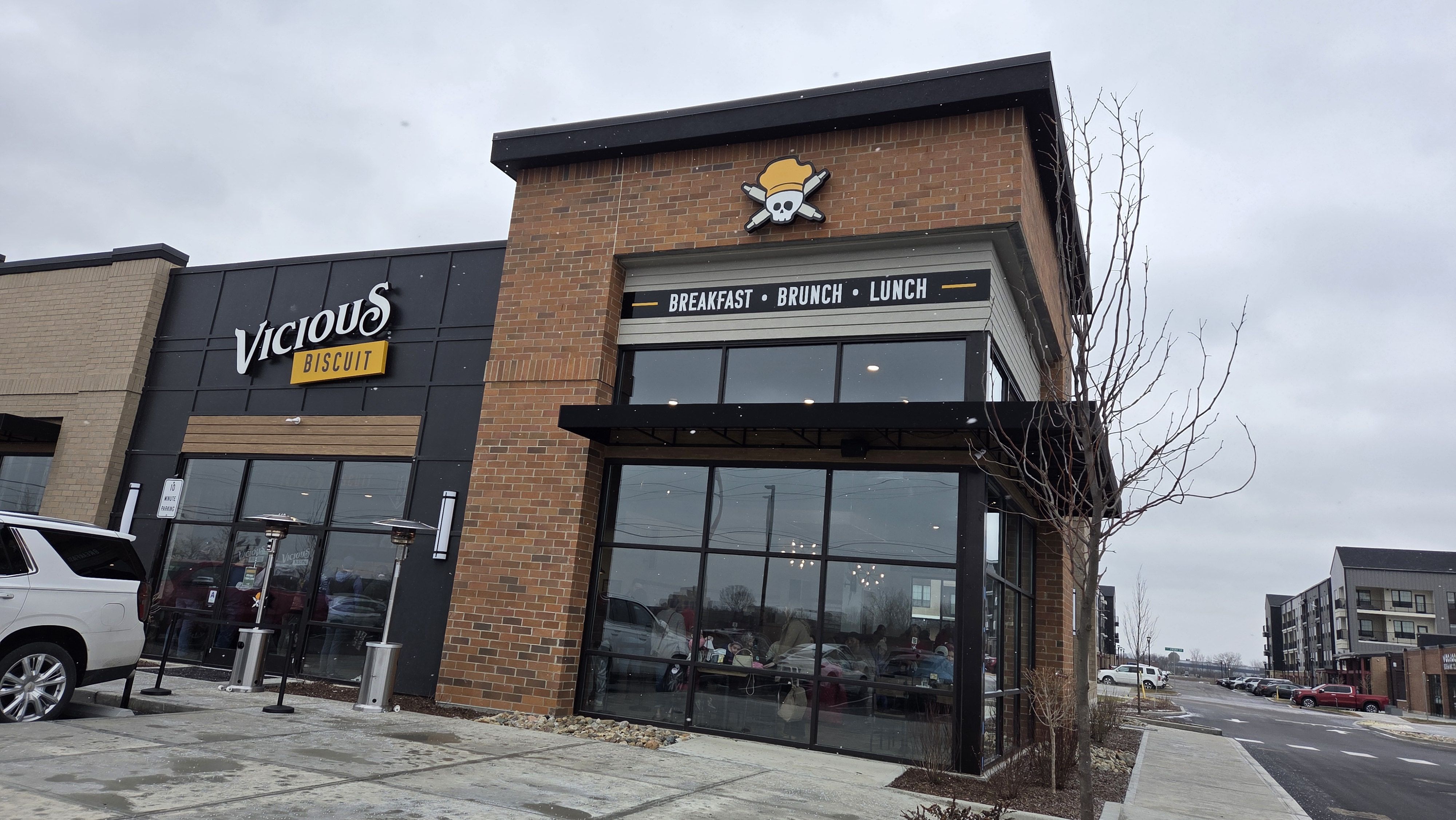 Exterior of Vicious Biscuit restaurant with brick and black facade, logo with skull and chef hat, signage reading "Breakfast Brunch Lunch," parked cars, overcast sky.