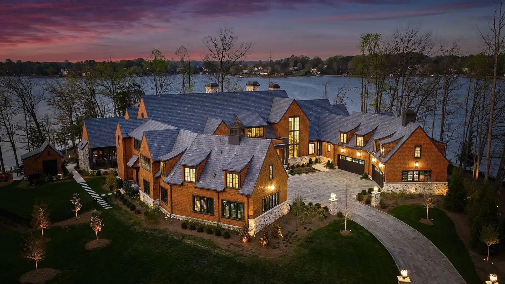 An expansive brick mansion with blue slate roofs sits lakeside at sunset. A curved stone driveway leads to the estate, with warm lights in the windows and a manicured lawn with young trees around.