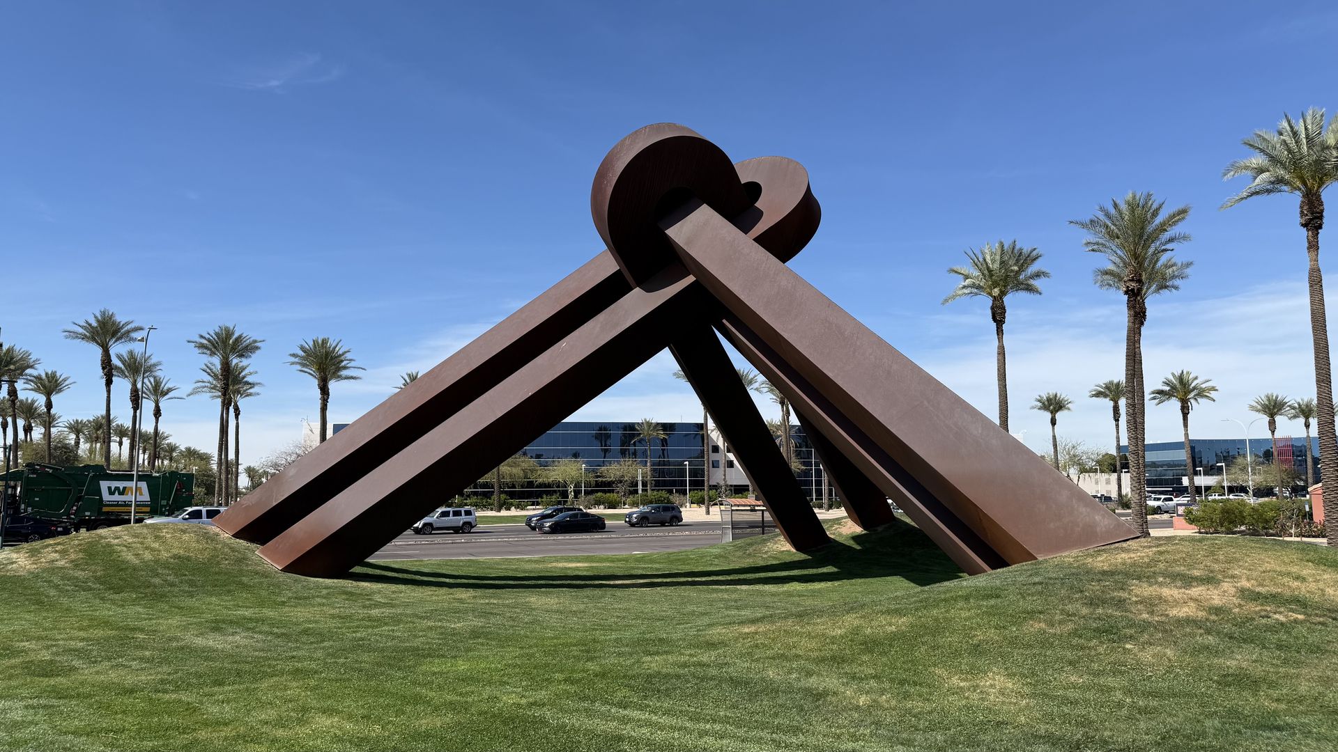 A large rust-colored metal sculpture of interlocking beams forming a loop, set on a grassy lawn with palm trees and a clear blue sky in the background; a street and buildings visible beyond.