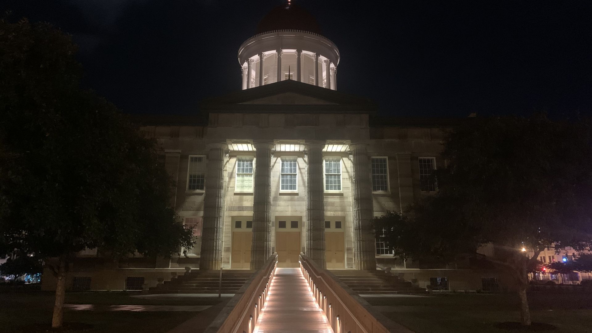 Night scene of a neoclassical building with a lit dome and flag atop. A bright walkway with railings leads to tall columns; dark sky and trees frame the facade.