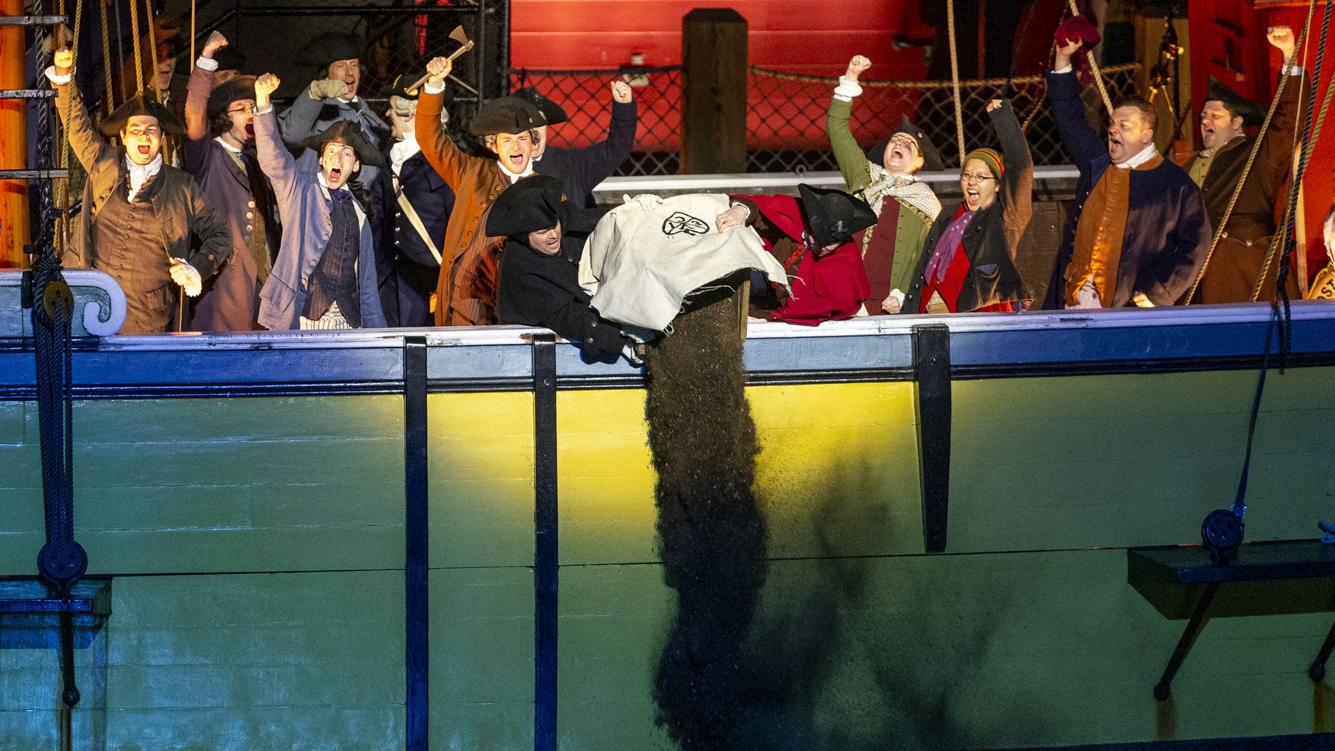 People in historical costumes on a green and yellow ship cheer as a man pours a sack of dark material off the side into the water at night.