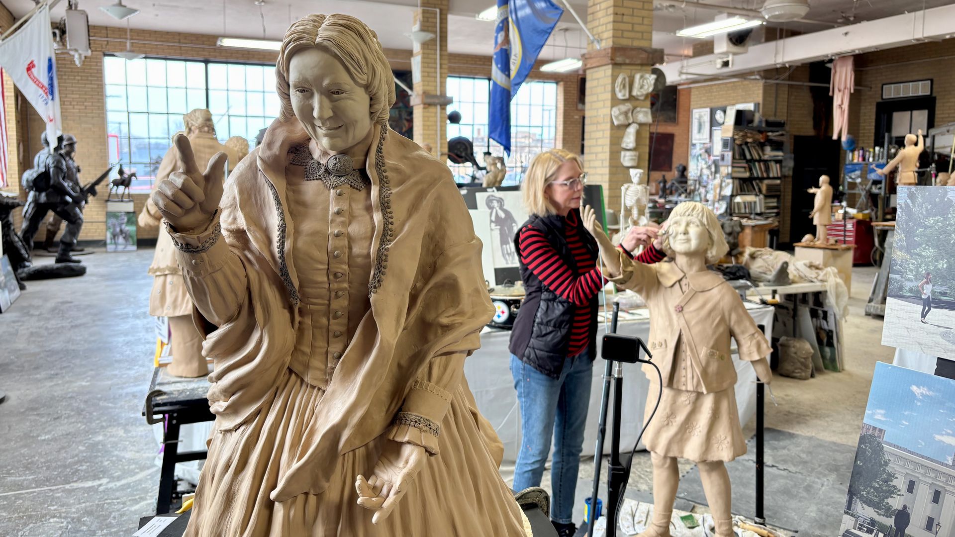 Bright sculpture studio with a life-size clay statue of a Victorian woman raising her finger; another artist shapes a smaller girl statue nearby, amid brick columns and flags.