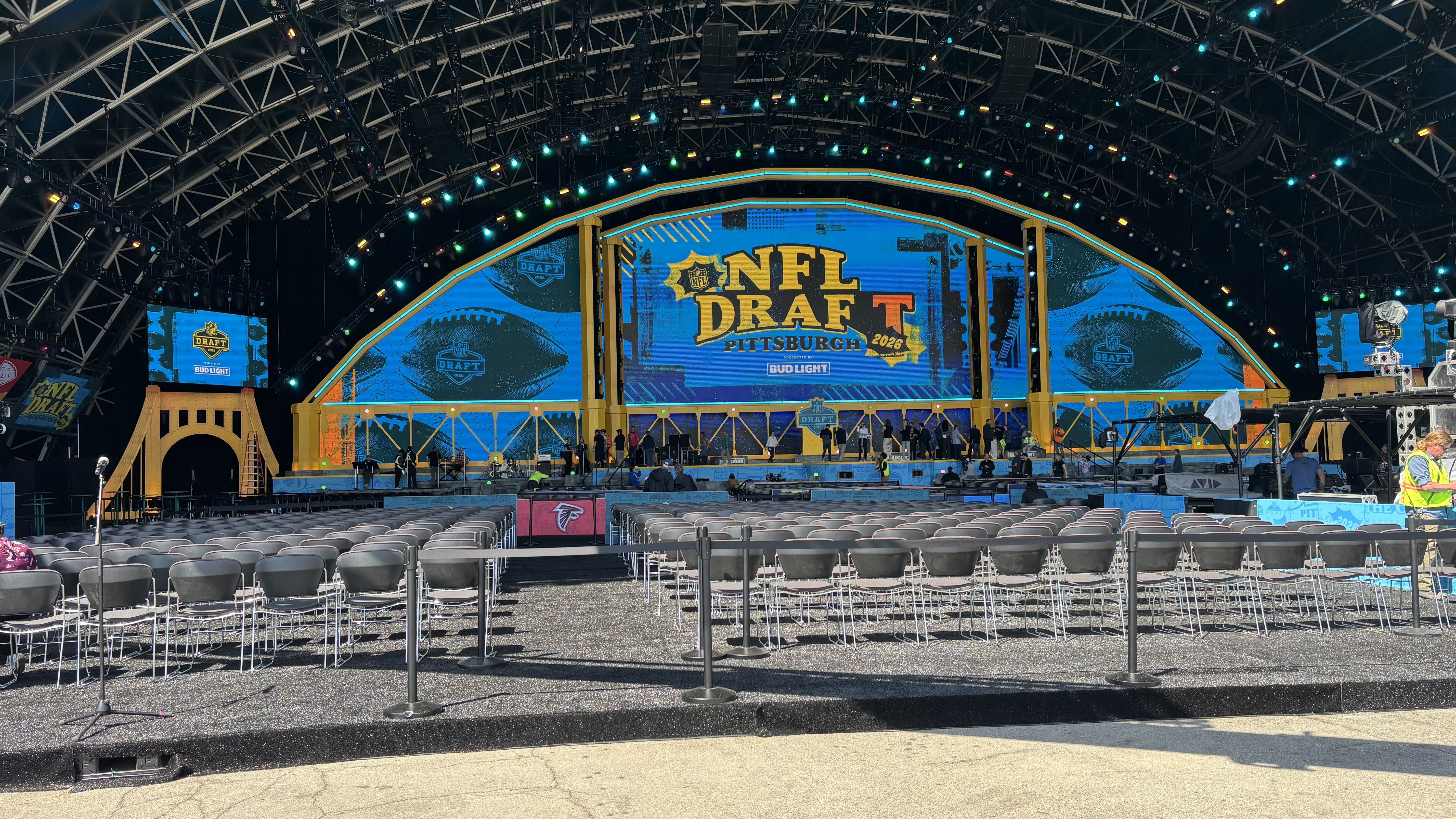 A large indoor stage for the NFL Draft in Pittsburgh, featuring a blue curved screen displaying "NFL DRAFT" and "PITTSBURGH"; colorful lights, overhead rigging, and rows of empty chairs in front.