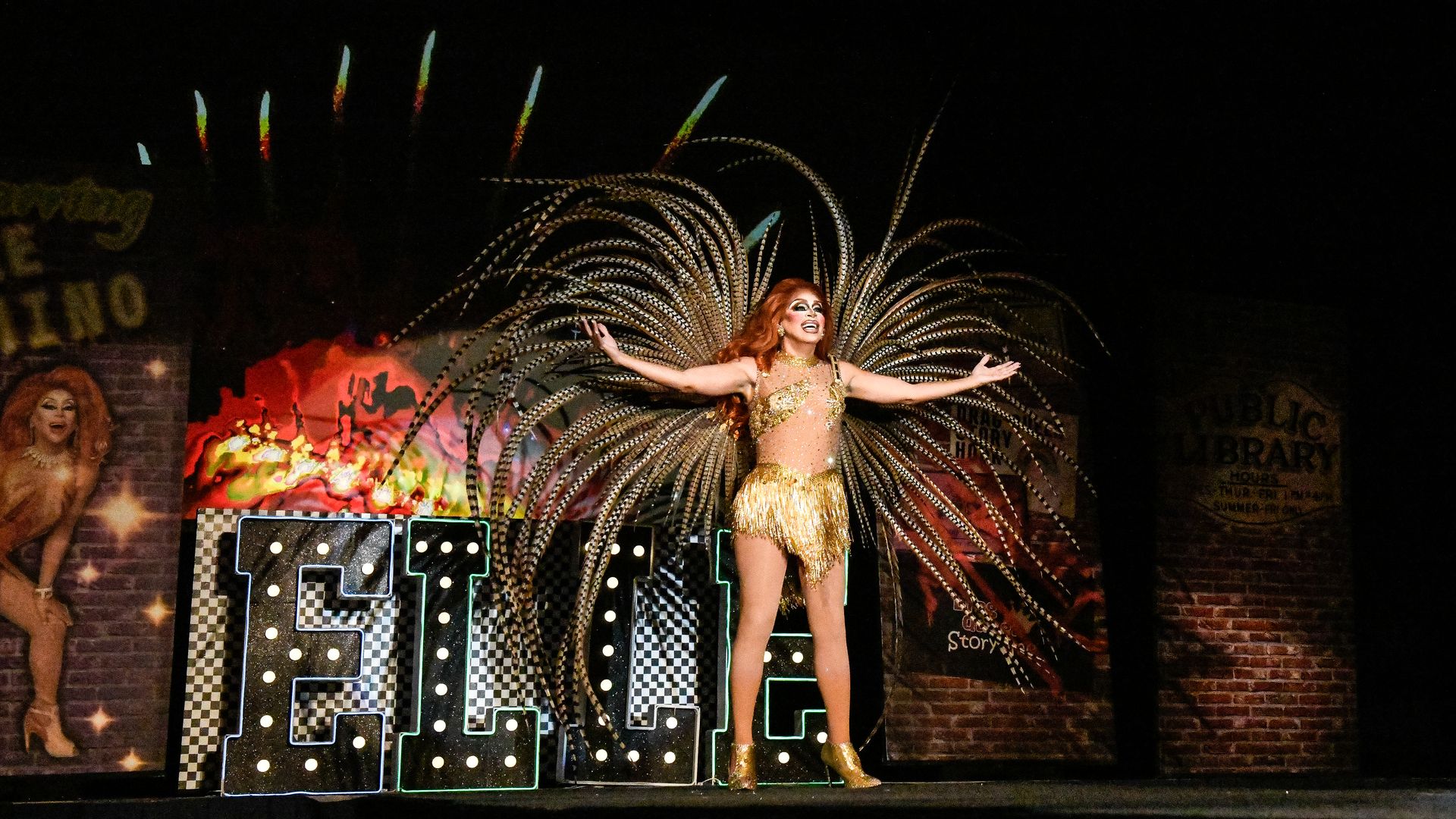 Showgirl in a gold sequined outfit wearing a massive silver feather headdress, arms outstretched on a brick-wall stage backdrop, with another performer visible on the left.