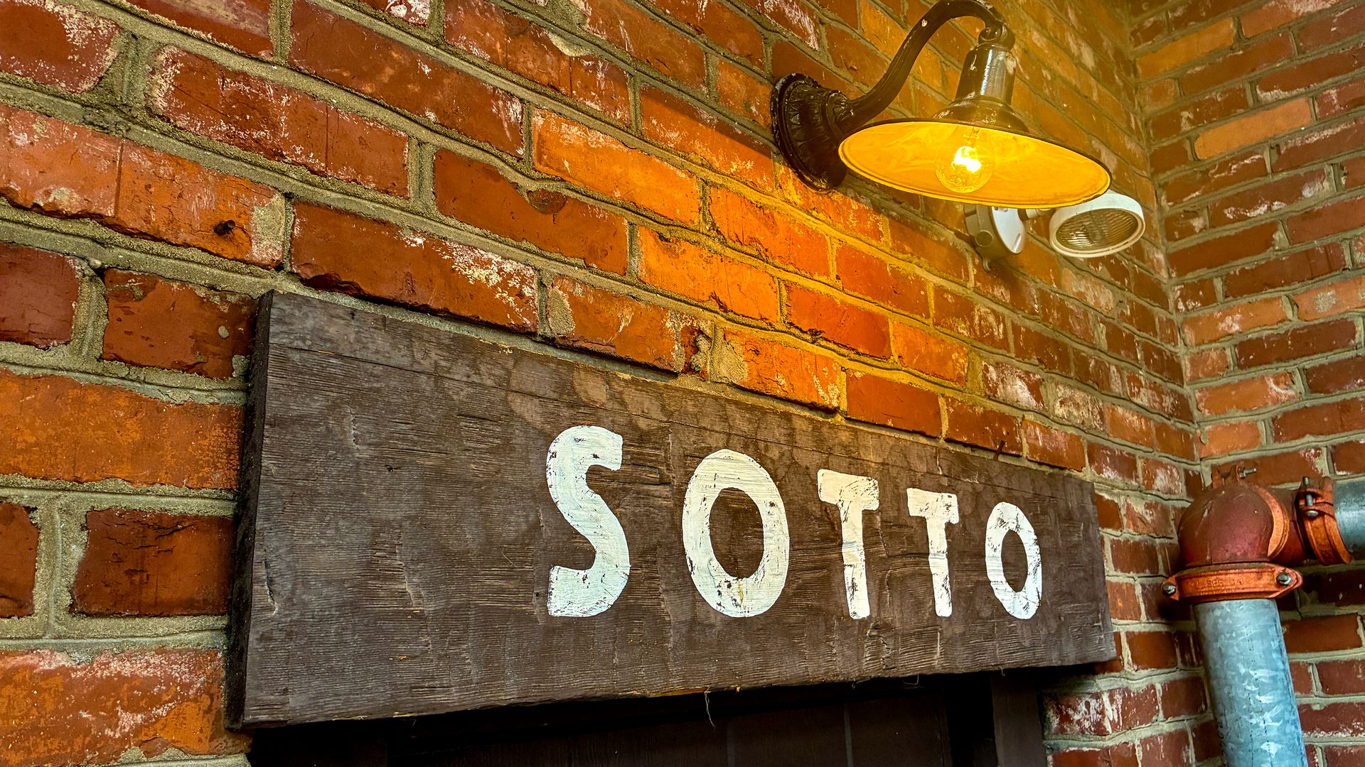 Amber outdoor wall light on a red brick wall, glowing warmly. A weathered dark wooden sign with large white letters hangs below, while a rusted pipe runs along the corner.