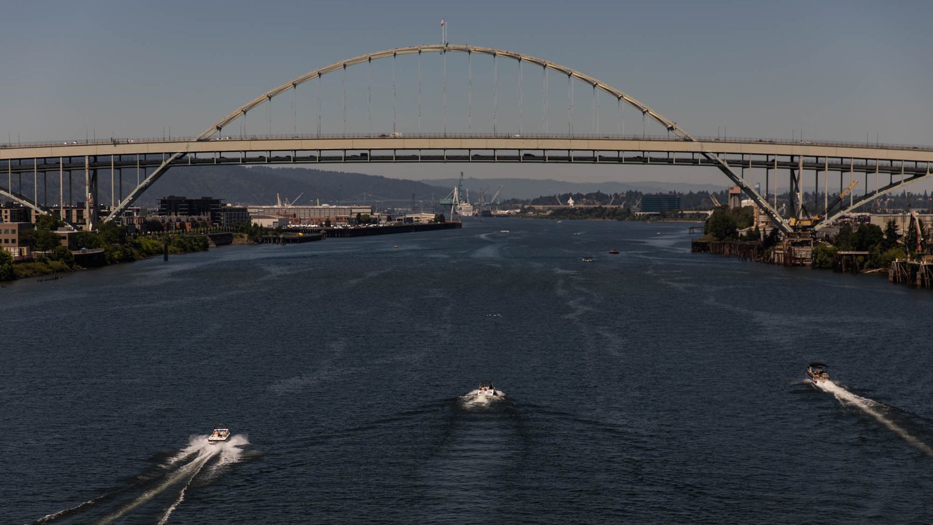 Wide river with a large arched bridge overhead. Three speedboats streak white wakes toward the foreground as city buildings line the banks and distant hills rise beyond.