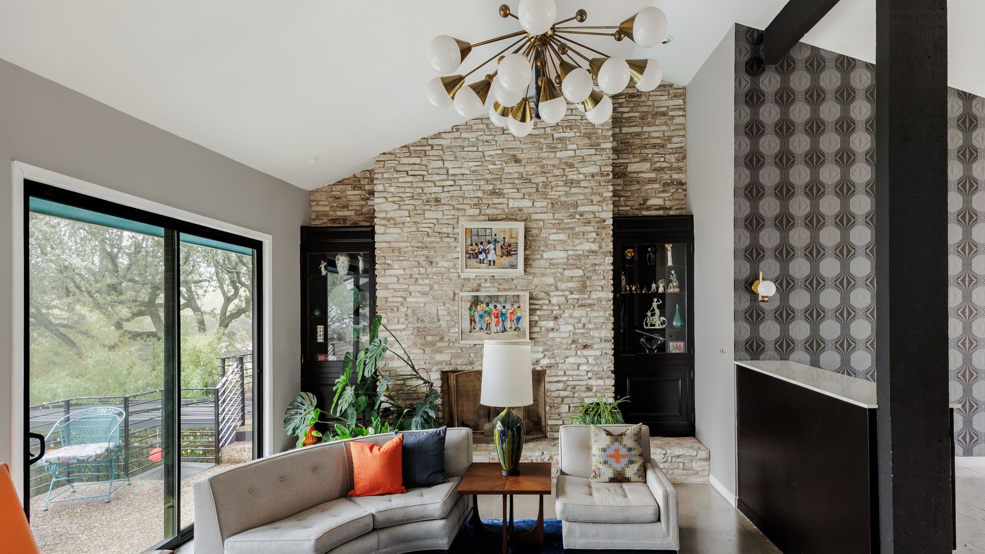 Living room with a tall stone fireplace, starburst chandelier, gray curved sofa with orange pillow, blue rug, plants, and black display cabinets. Sliding glass door leads to a patio.