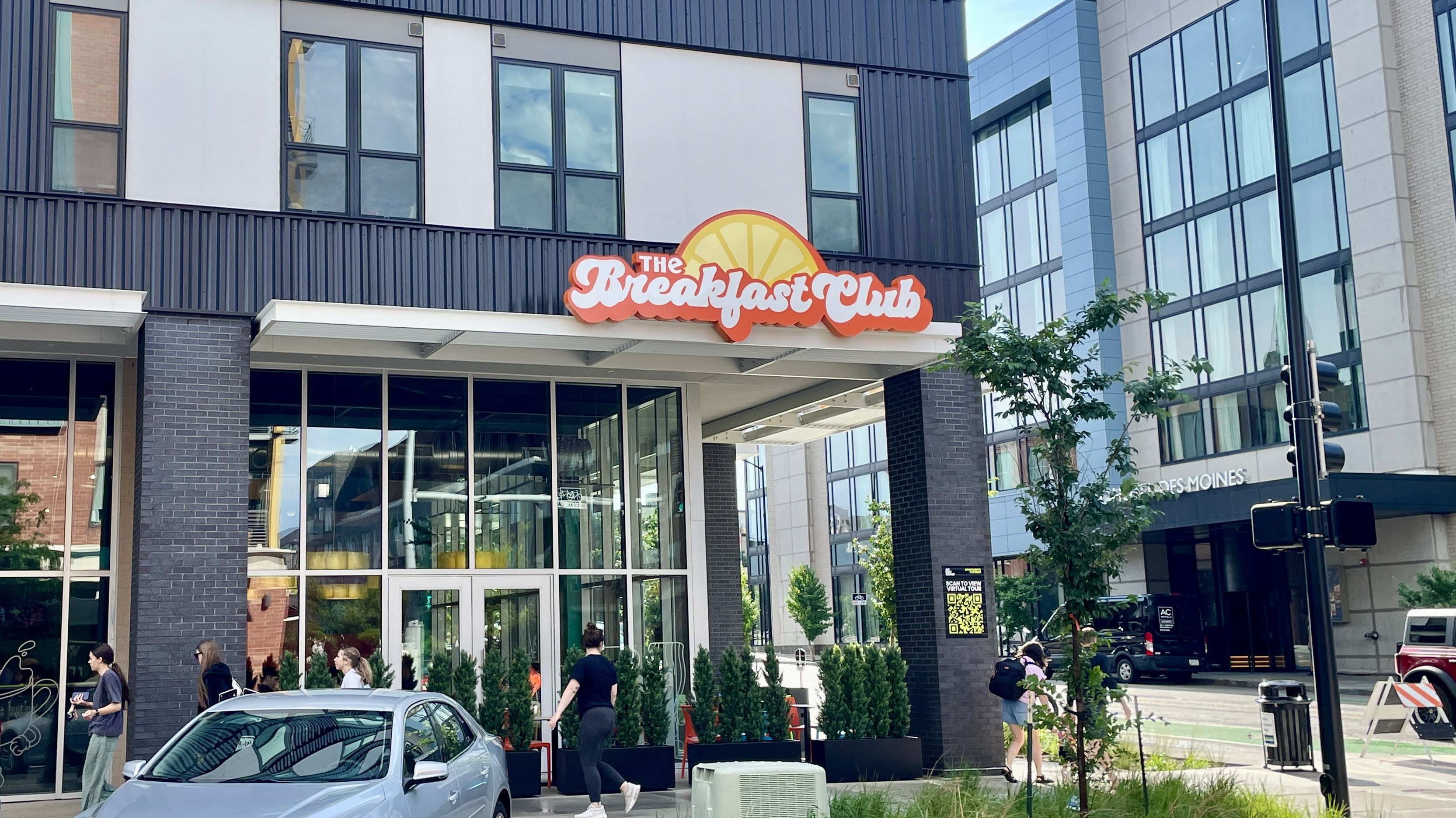 Urban street scene with a modern building featuring large windows and a bright orange sign reading "The Breakfast Club" with a sun graphic. Pedestrians pass by and a silver car is parked in front.