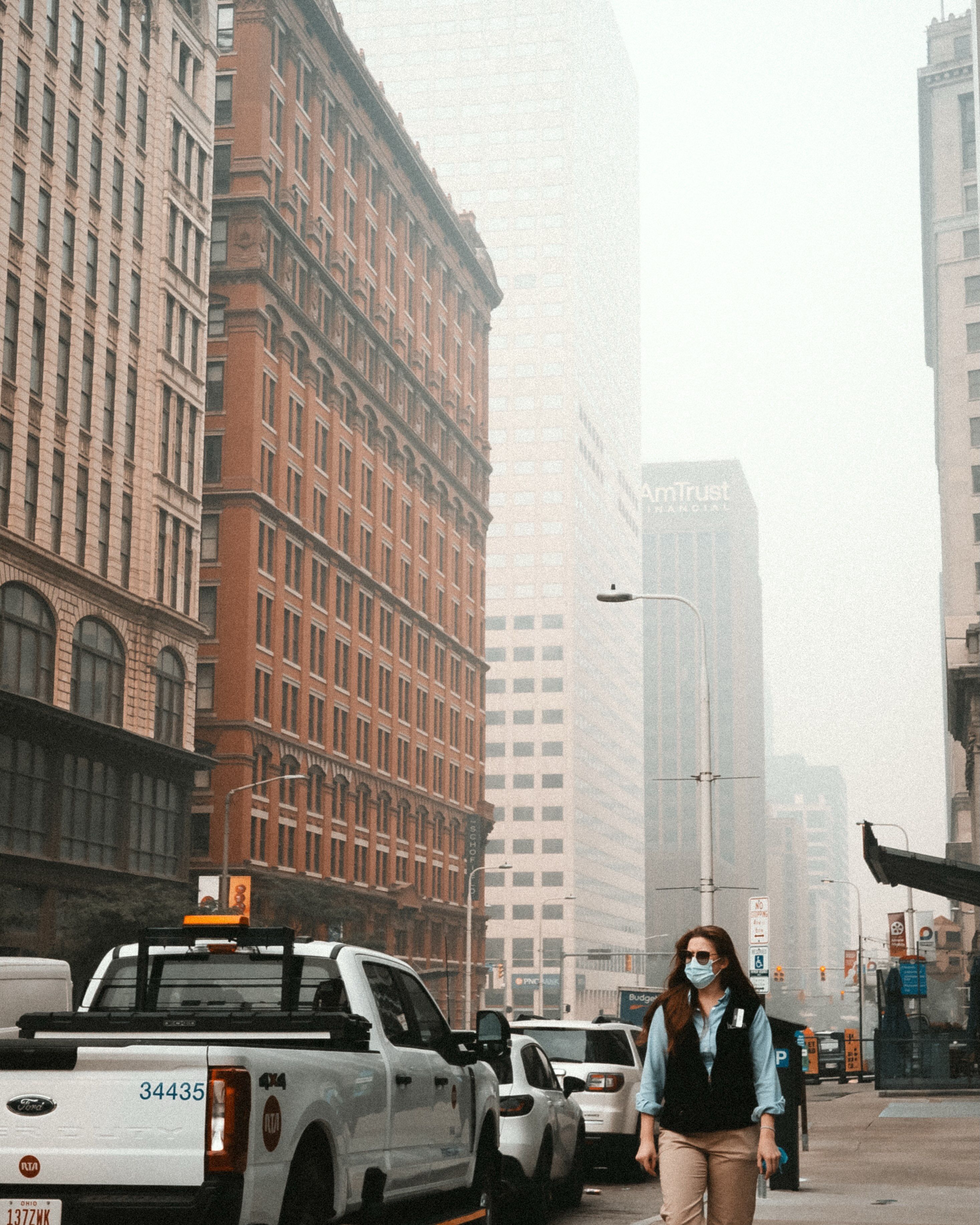 City street with red-brick buildings and a hazy skyline. A woman wearing a mask and a blue shirt walks past white delivery trucks; distant office towers fade in the fog.