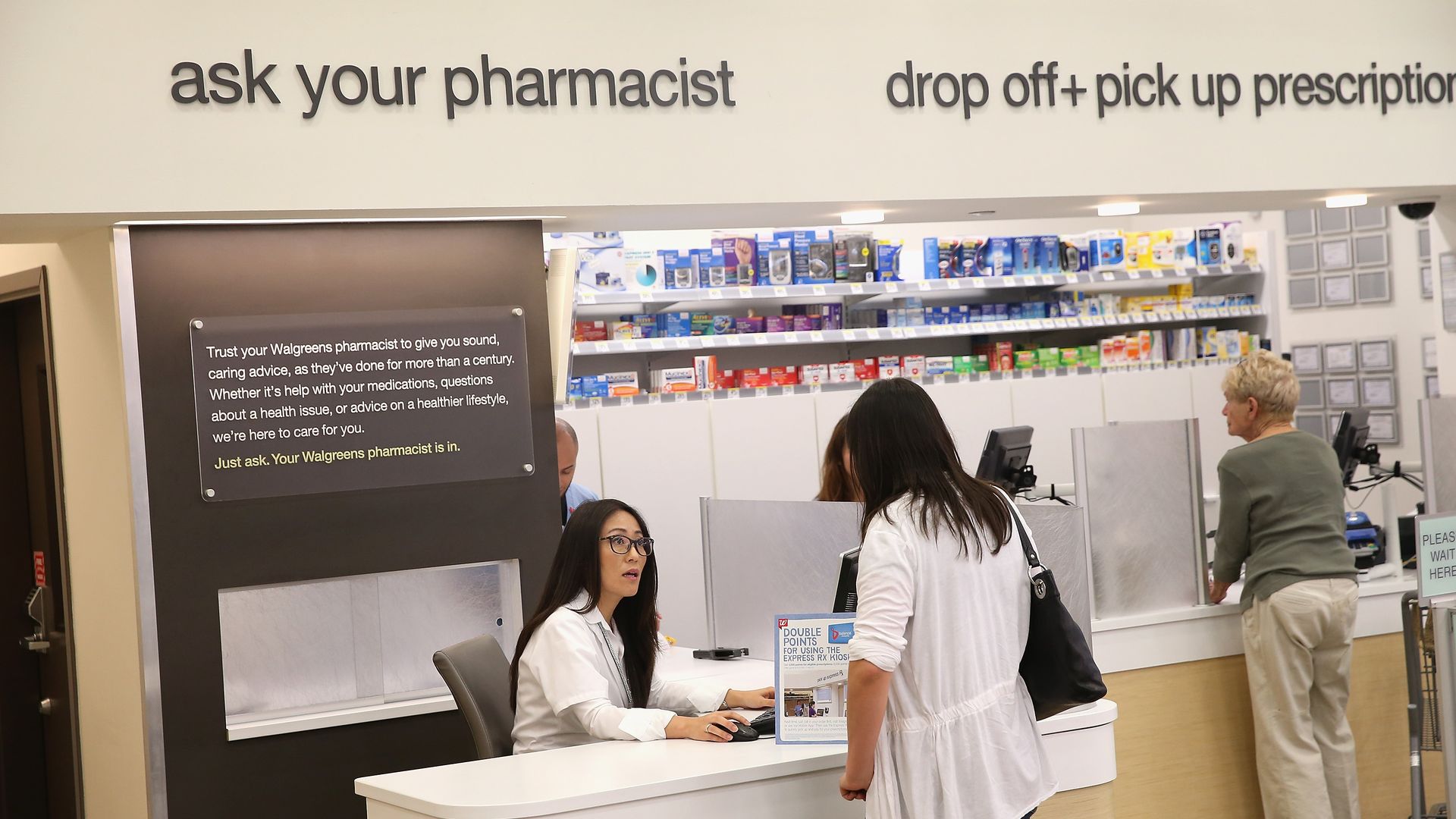 A pharmacists talks to a customer, who is standing in front of a counter.