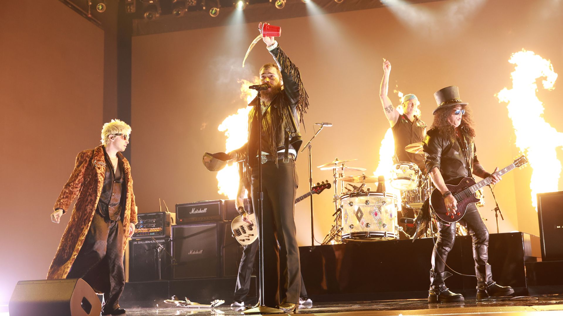 Rock band performs on stage with bright yellow flames behind them. Lead singer in black leather jacket pours liquid from red cup. Guitarist in top hat and sunglasses plays guitar