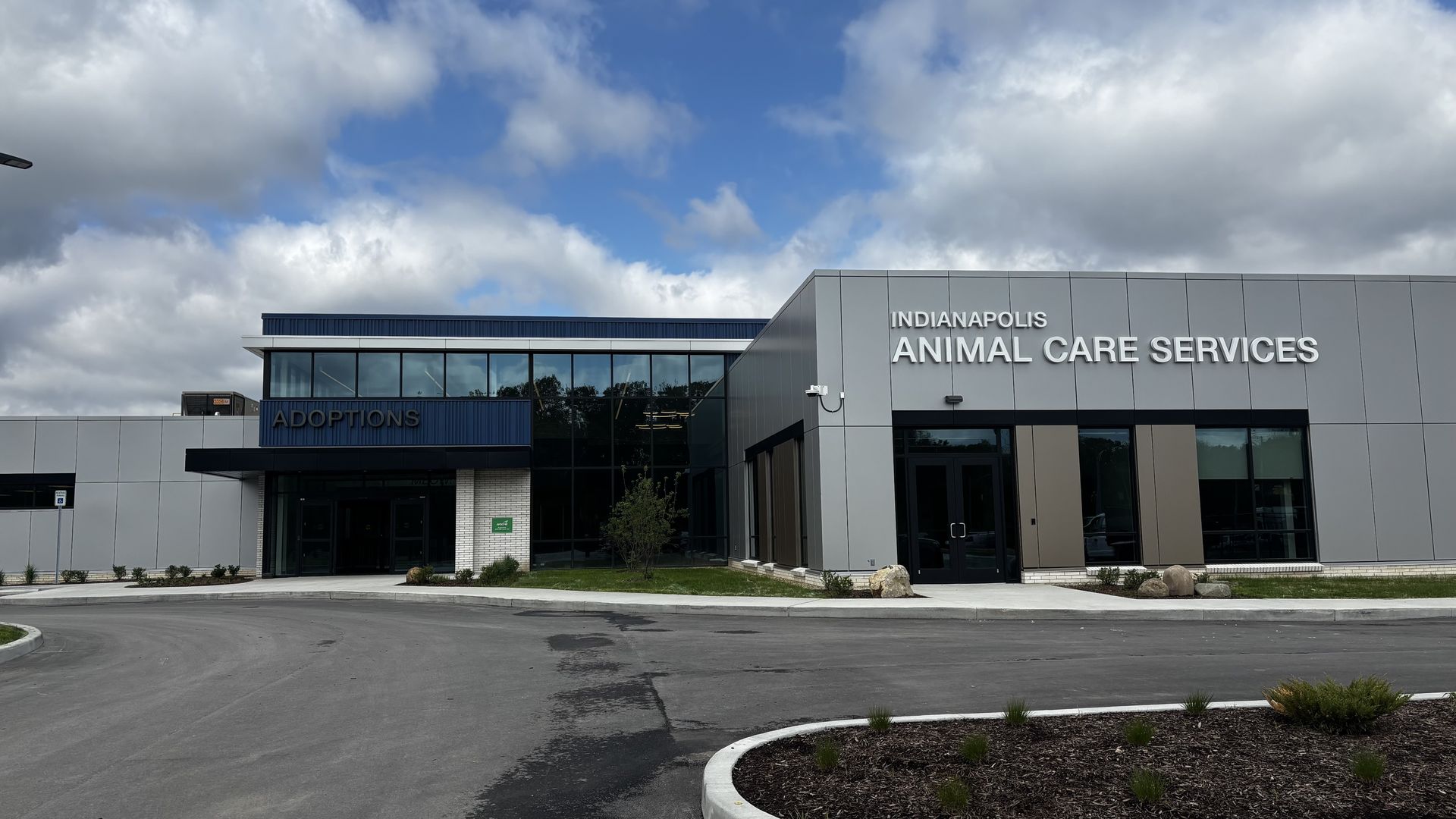 Exterior of the Indianapolis Animal Care Services building featuring a large glass facade, grey panels, and an ADOPTIONS sign; cloudy sky, curved driveway, and small landscaped beds in front.
