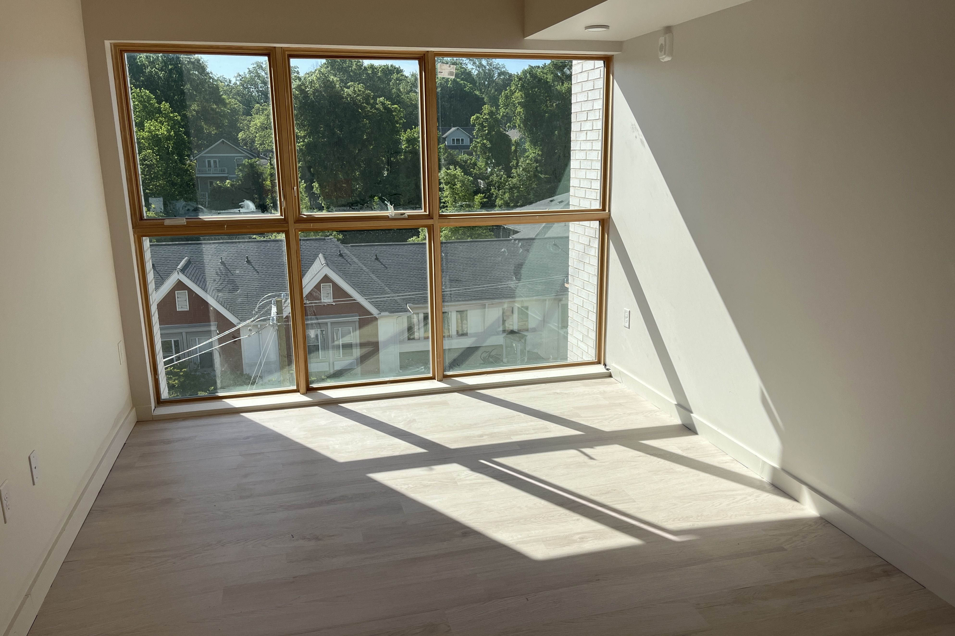 Bright empty room with a large wood-framed window, sunlight casting long shadows on light oak floors. Outside, green trees and rooftops are visible; the right wall has a brick column.