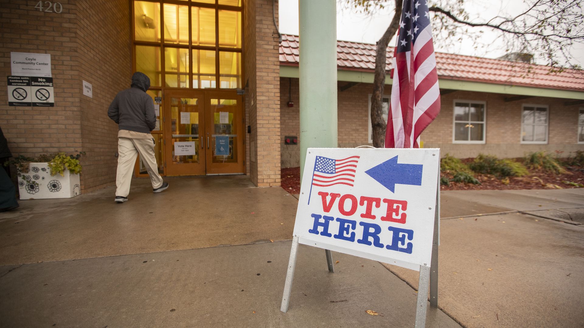 A vote here sign with an arrow and an american flag, poining into a building with a man walking in.