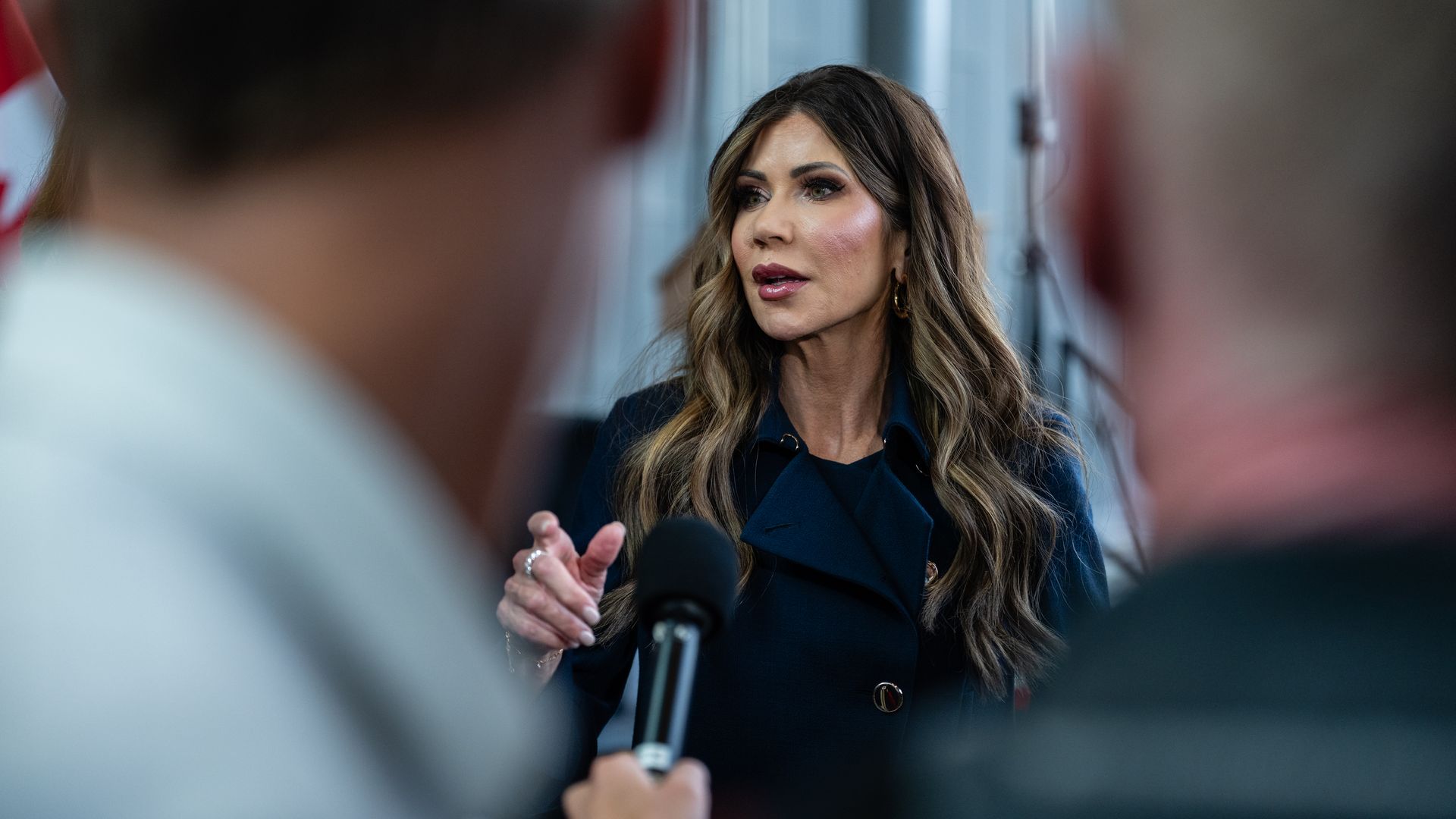 Woman with long brunette hair and gold hoop earrings speaks into a microphone held by a person in the foreground, wearing a dark blue coat, with a blurred indoor background.