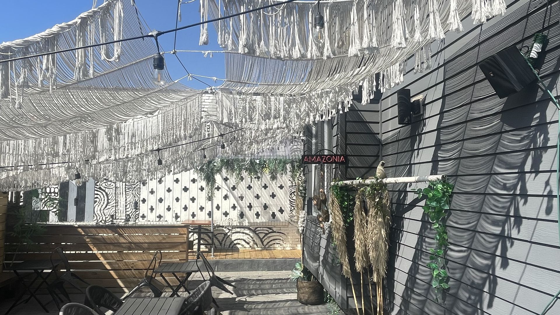 Outdoor patio with black tables and chairs, wooden fence, patterned white walls, hanging string lights, white fringe canopy, and green plants under a clear blue sky.