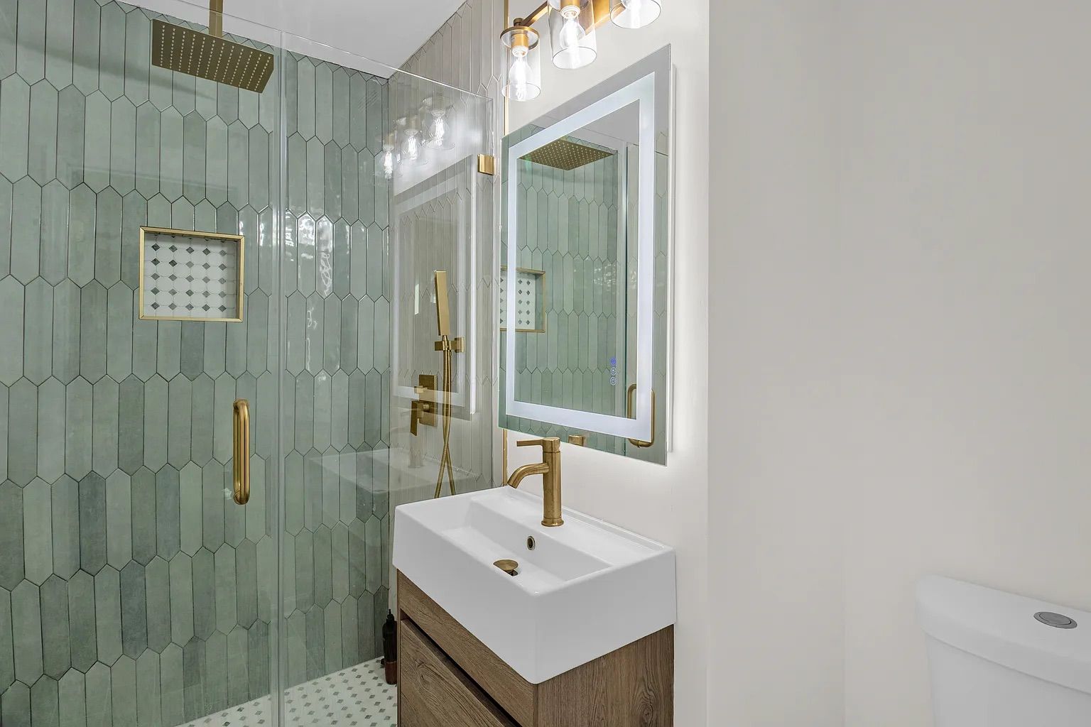 Modern bathroom with glass shower featuring green hexagonal tiles, gold fixtures, a white sink with gold faucet, illuminated rectangular mirror, and white walls.