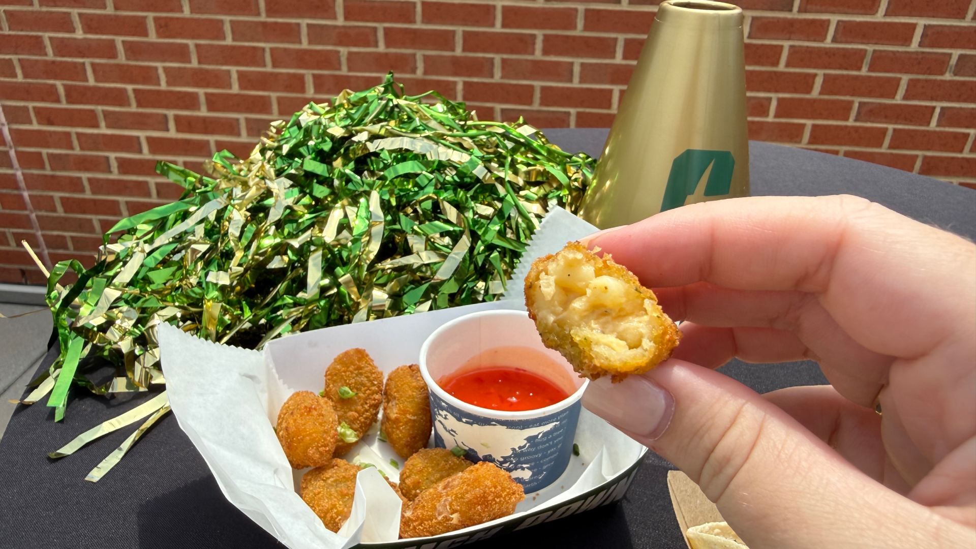 Close-up of a hand holding a crispy breaded bite over a tray of fried snacks with marinara dipping sauce; green-and-gold tinsel and a gold party cone sit on a table, brick wall in the background.
