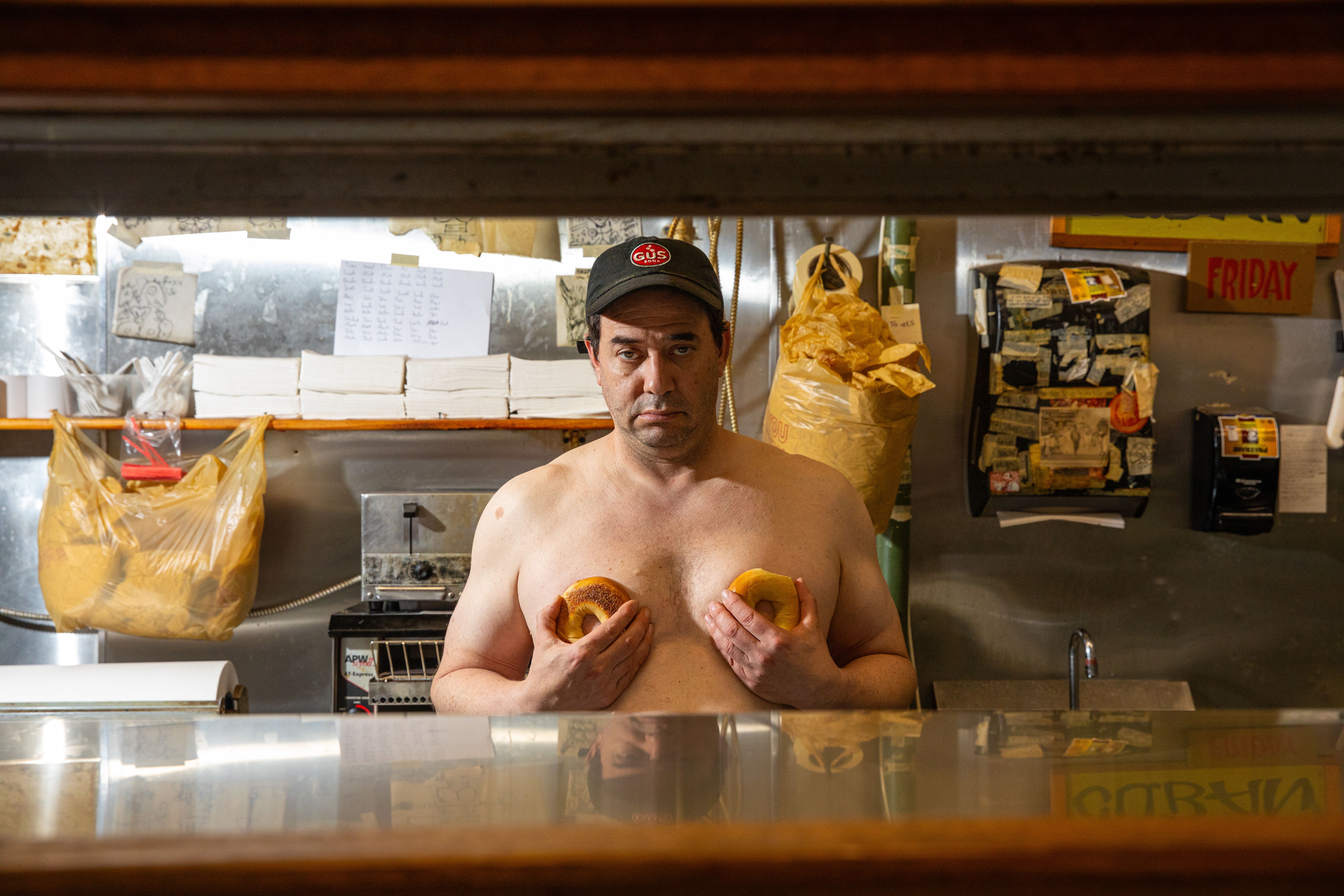 A man stands behind a deli counter, shirtless, with two bagels help in front of his chest.