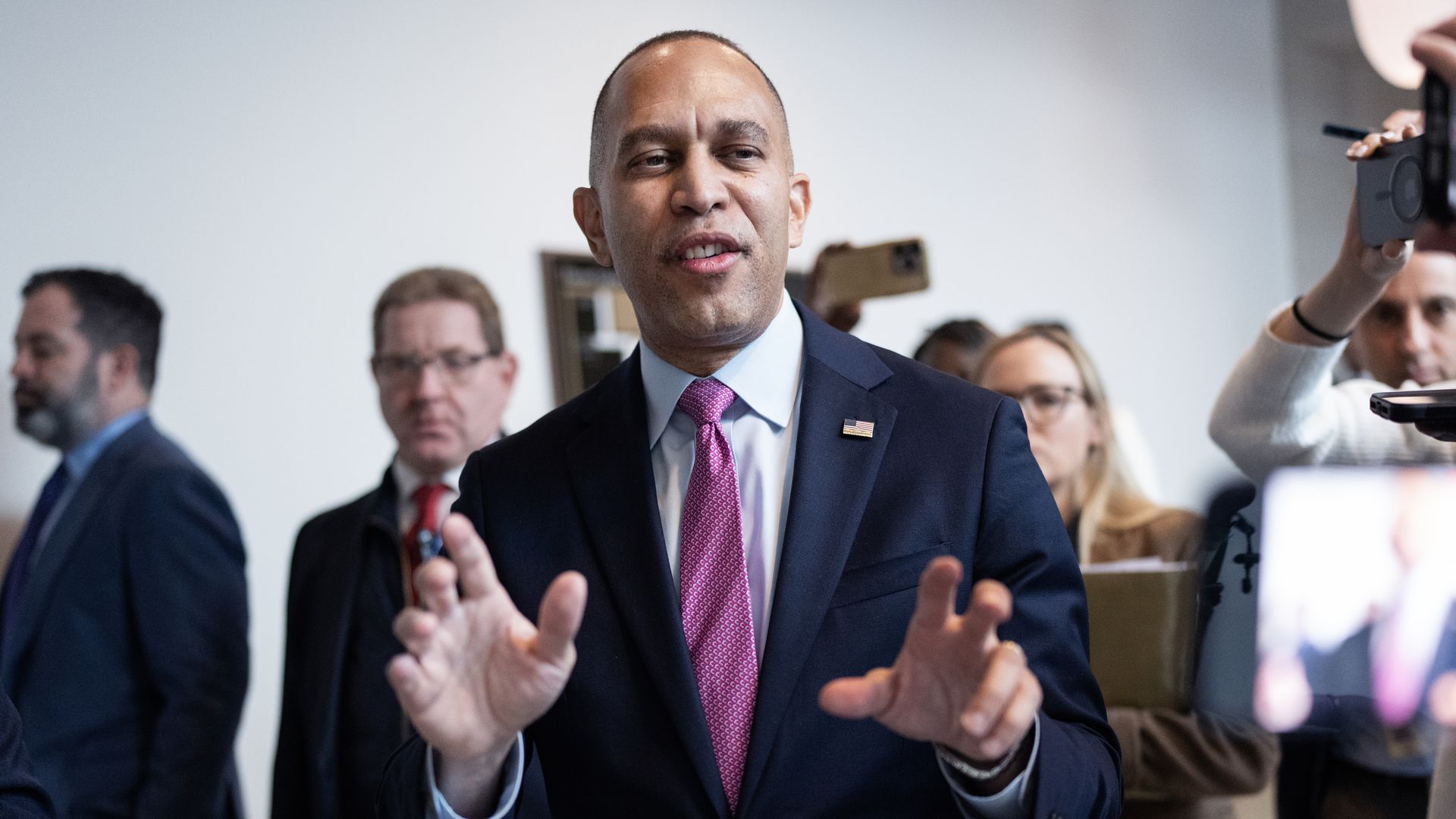 House Minority Leader Hakeem Jeffries, wearing a blue suit and lifting his arms while flanked by staff and reporters.