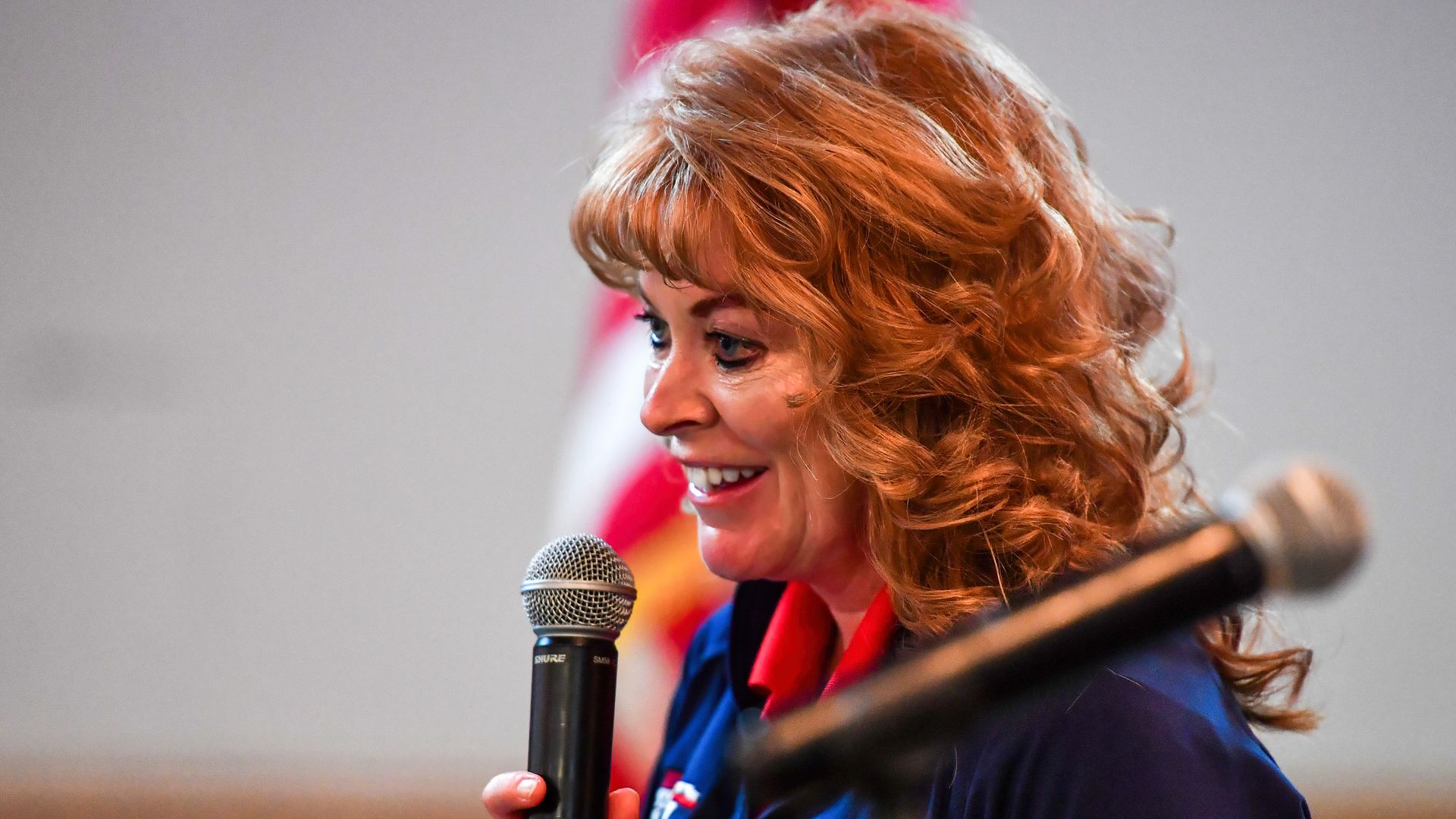 Woman with curly red hair wearing a navy blue jacket holding a microphone, smiling while speaking, with an American flag blurred in the background.