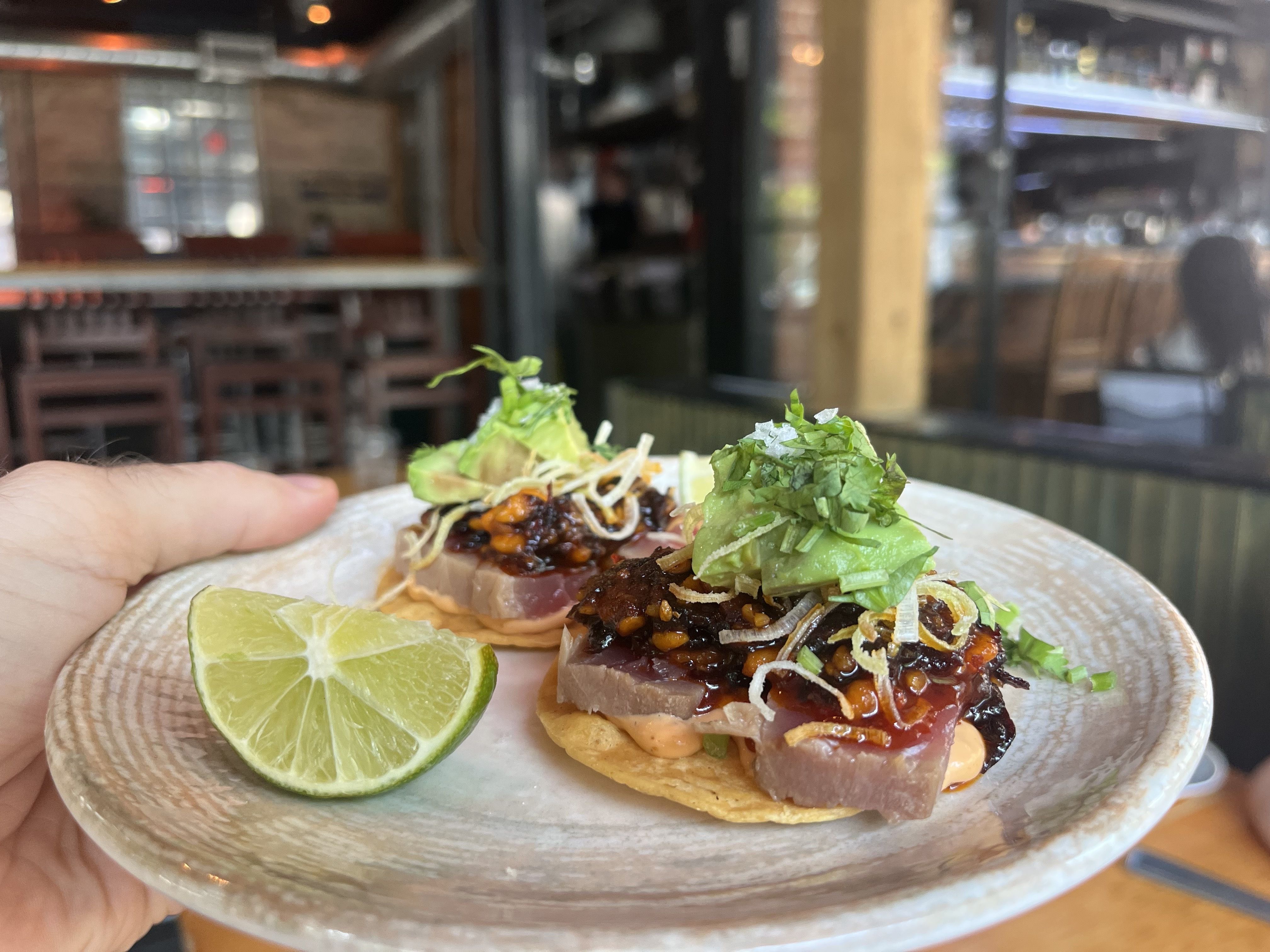Close-up of two open-faced tacos on a plate held by a hand, topped with dark sauce, cilantro, onions, cheese, and lime; blurred restaurant interior in the background.
