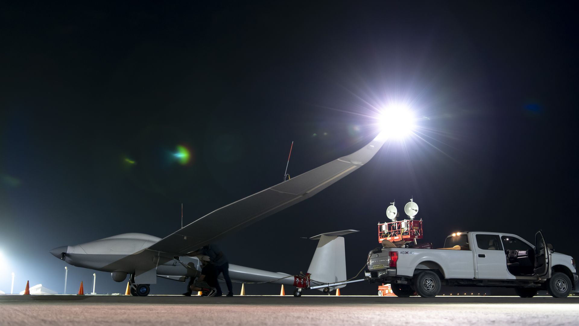 A large military drone sits on the tarmac near a white truck. The sky is black. The wingtip is brightly illuminated.