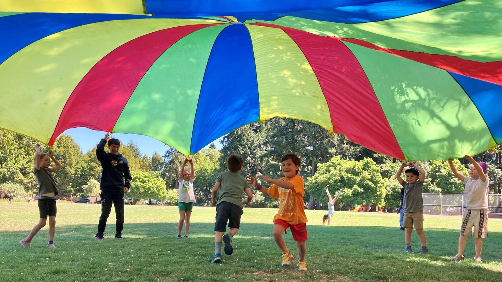 Children and a caregiver playing under a colored parachute at at summer camp in a Portland park.