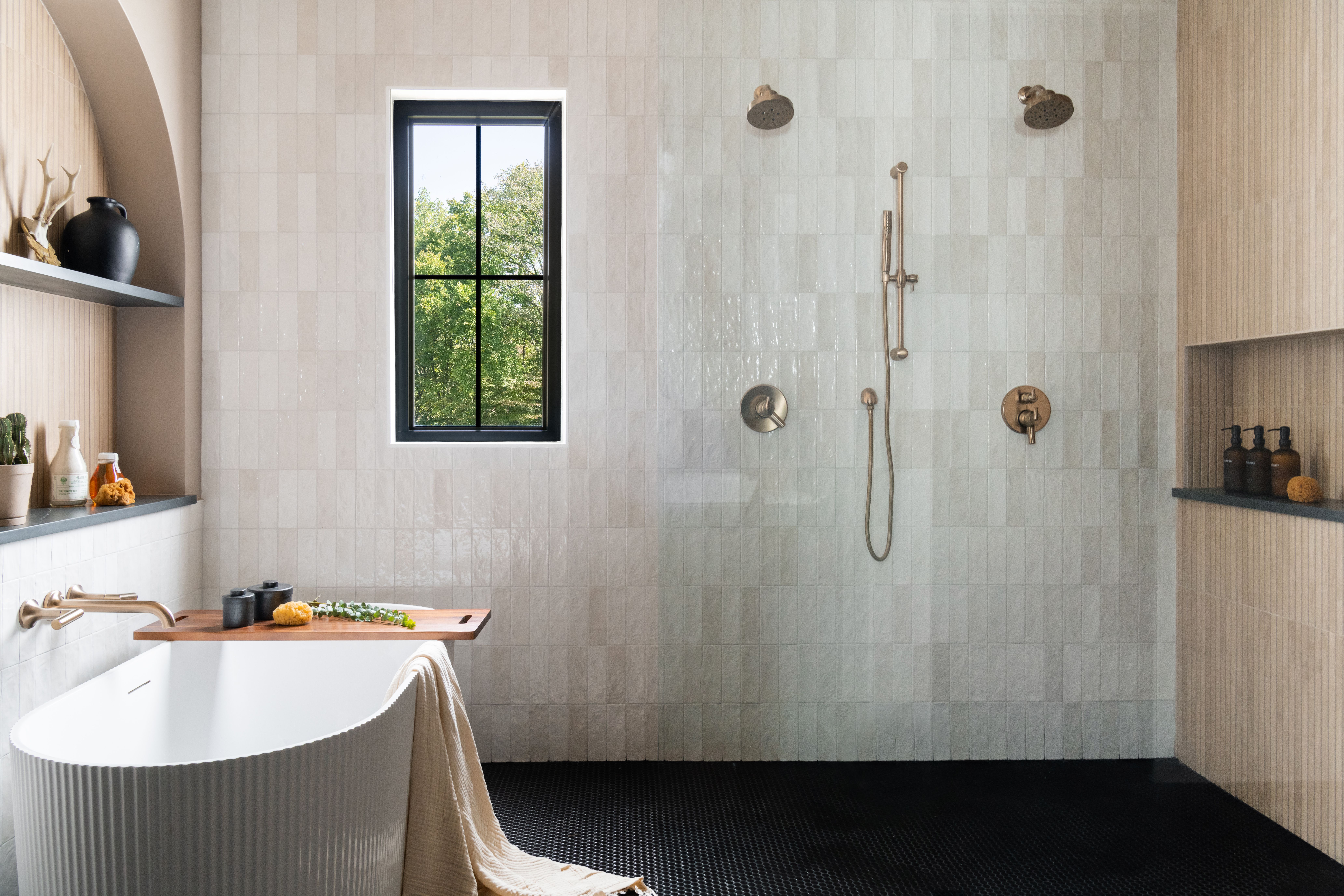 Modern bathroom with white freestanding tub, black hex tile floor, dual showerheads and wooden shelves with decor and toiletries. A frameless, floor-to-ceiling glass partition sits between the tub and shower area.