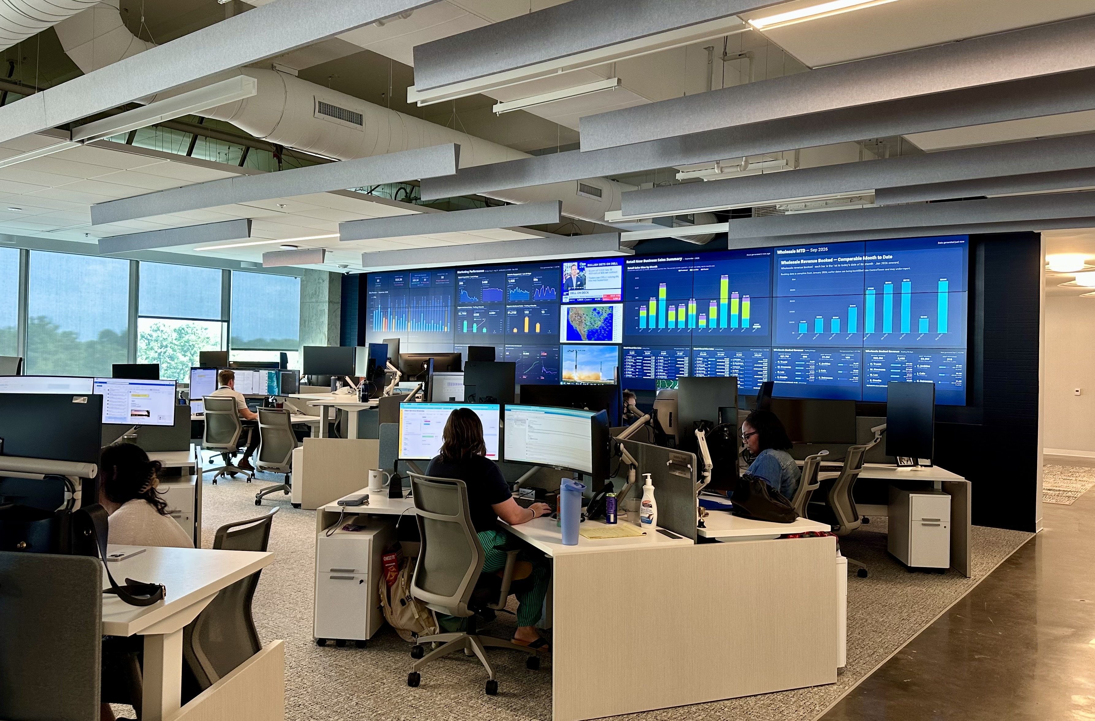 Open-plan trading floor with rows of desks and computer monitors, a wall of blue data dashboards, and suspended gray acoustic panels overhead; daylight enters from large windows on the left.