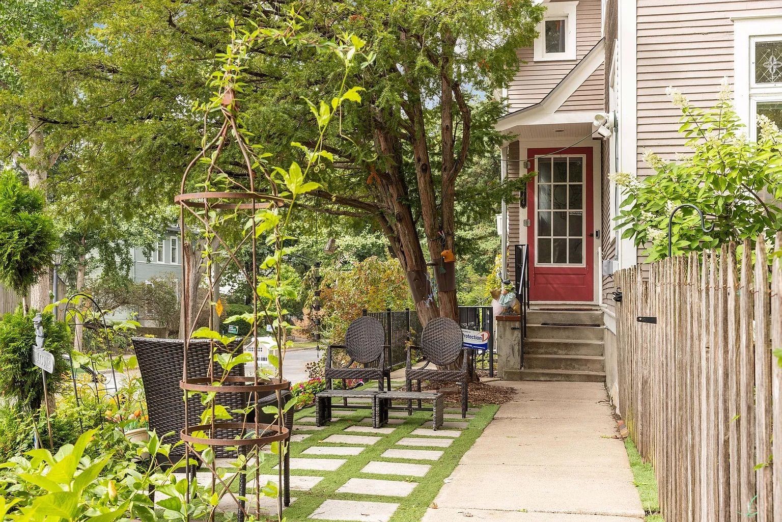 Outdoor patio with dark brown wicker chairs and a small table on stone and grass pathway beside a beige house with red door and wooden fence, surrounded by green trees and plants.