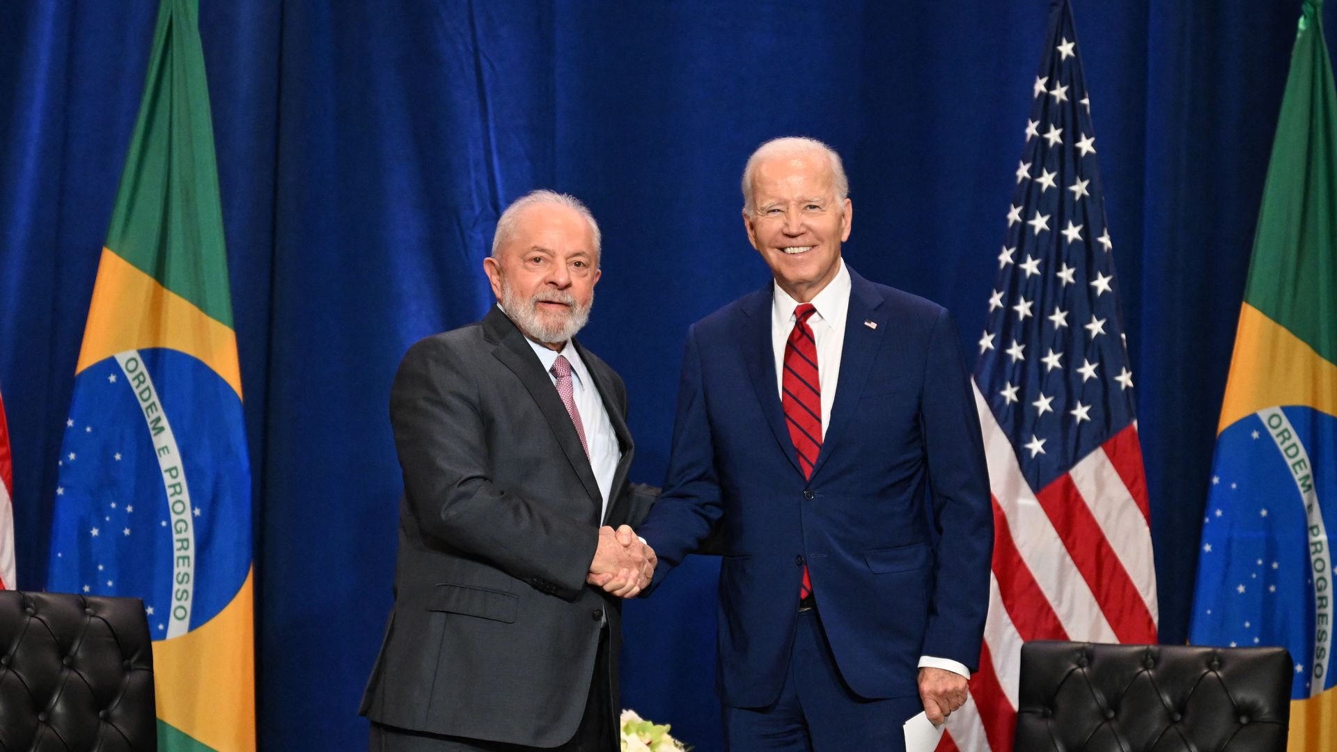 Brazil president Lula stands to the left of President Joe Biden, shaking hands. the Brazil and American flags are behind them as they stand in front of a blue wall