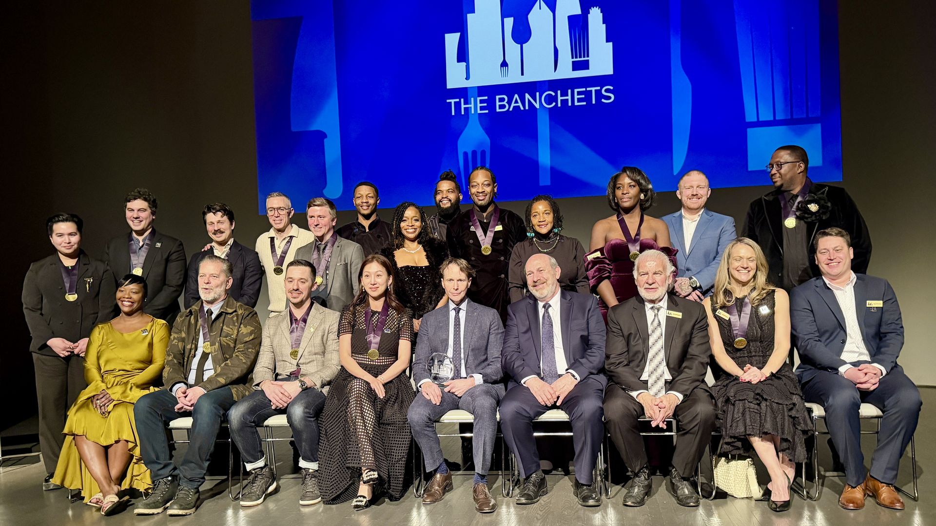 Group photo of 21 diverse people dressed formally, some wearing medals, seated and standing on stage with a blue backdrop displaying "THE BANCHETS" and city skyline graphic.